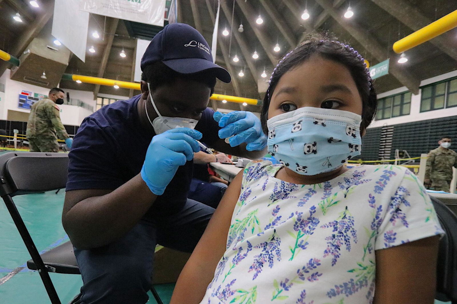 FEMA emergency medical technician Shane Alexander, left, prepares to administer the first dose of a Covid-19 vaccine to 6-year-old Elaina Guzman, as she looks on during a vaccination clinic held Thursday at the University of Guam Calvo Field House.