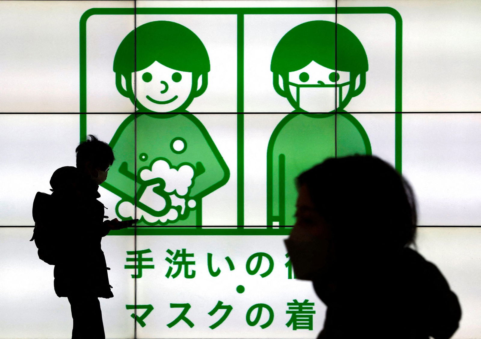 Passersby wearing protective face masks walk past in front of an electric screen displaying notice about Covid-19 safety measures in Tokyo, Japan, Feb. 1, 2022.