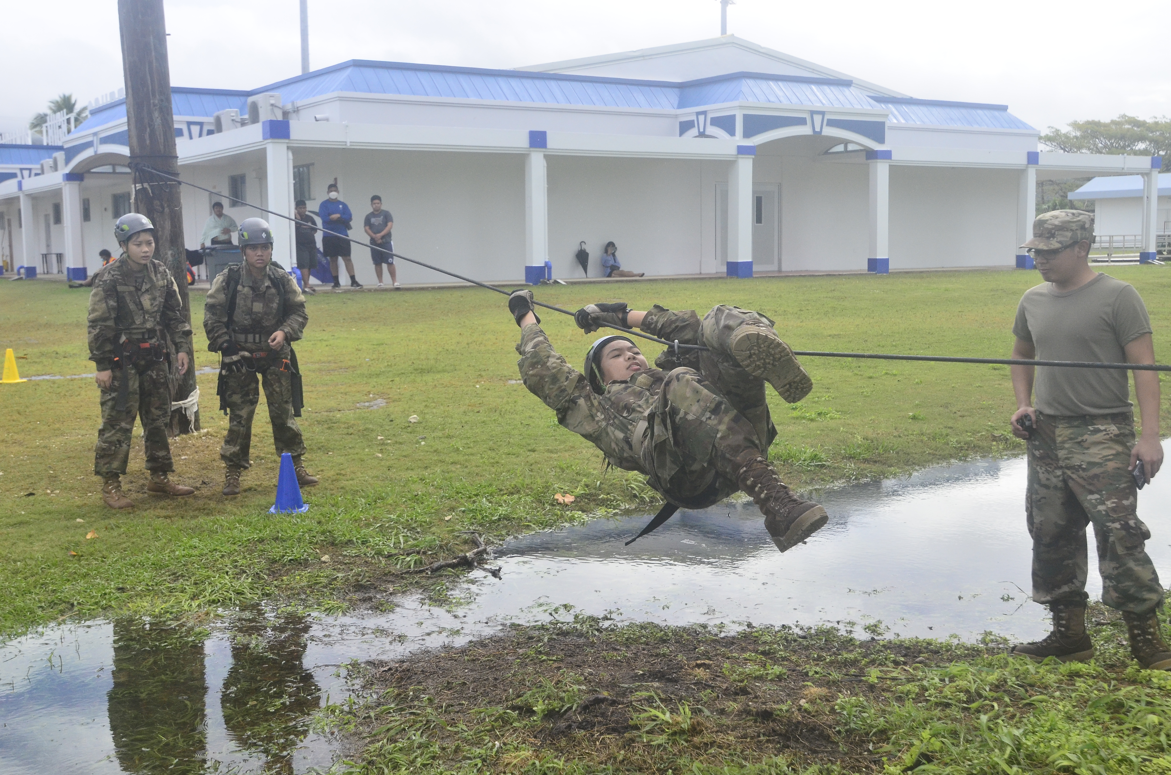 A cadet executes the Rope Bridge obstacle as her teammates look on.