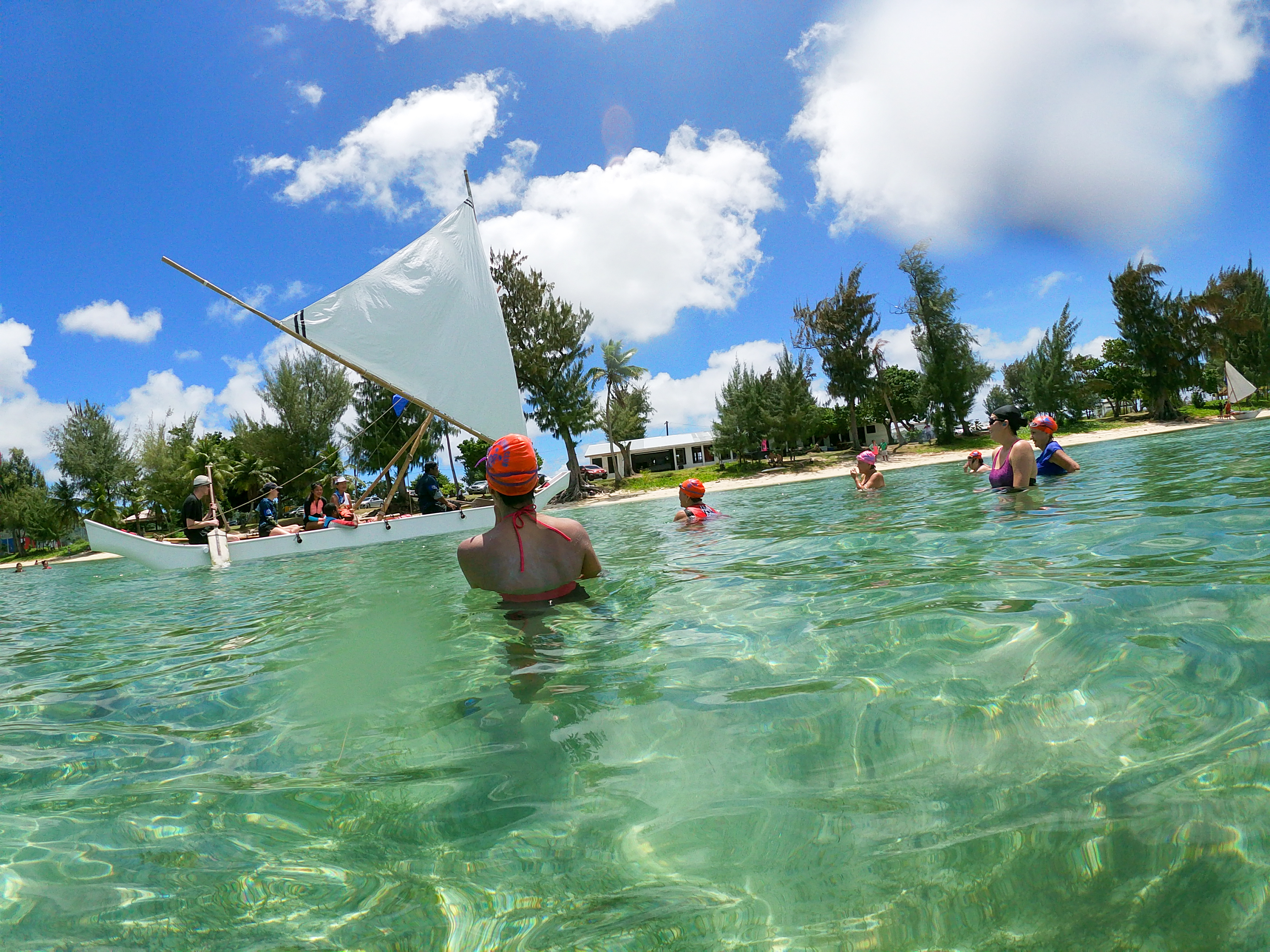 Photo shows Sirena Project swimmers as Neni, a white sail canoe, sails by the beach off Guma Sakman.