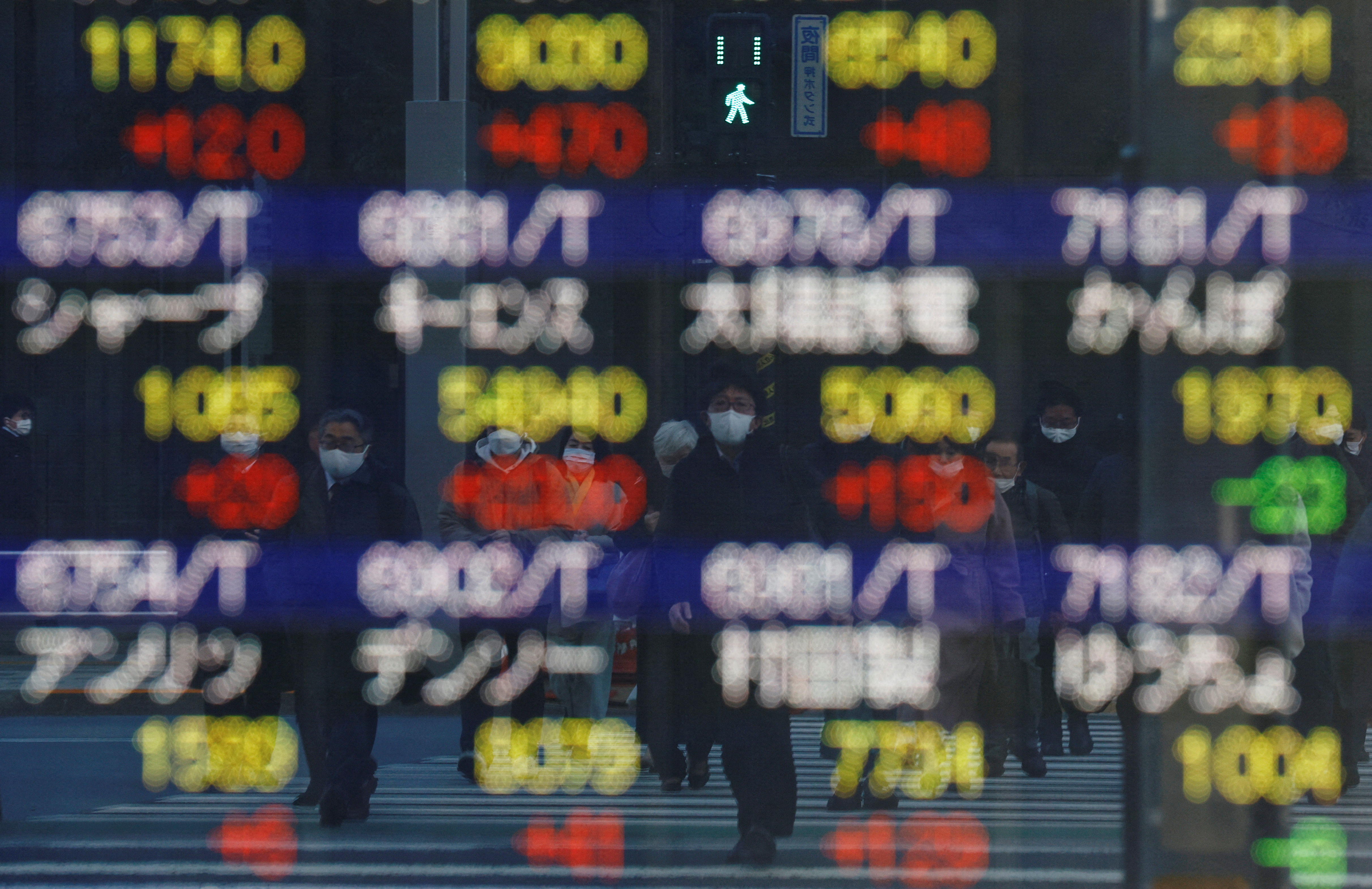 Pedestrians wearing protective masks are reflected on an electronic board displaying various company’s stock prices outside a brokerage in Tokyo, Japan, Feb. 25, 2022.