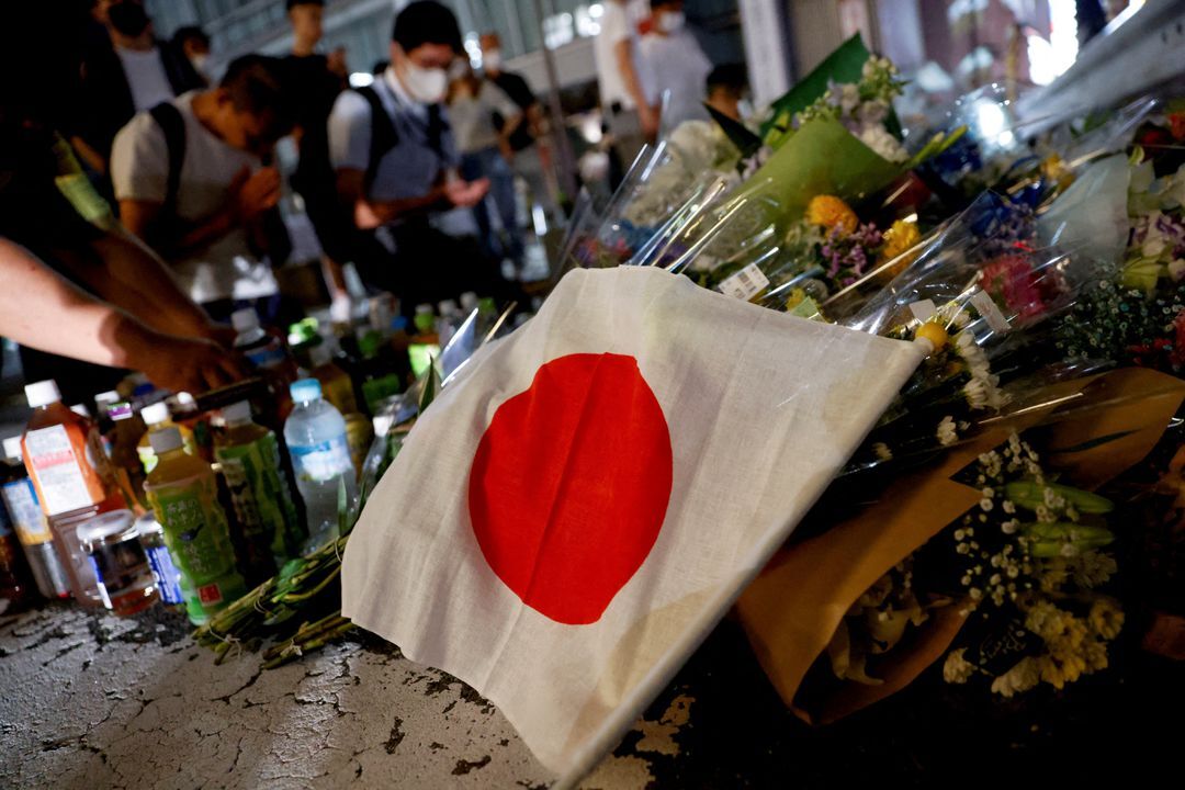 A Japanese flag is seen as people pray next to tributes laid at the site where late former Japanese Prime Minister Shinzo Abe was shot while campaigning for a parliamentary election, near Yamato-Saidaiji station in Nara, western Japan, July 8, 2022.