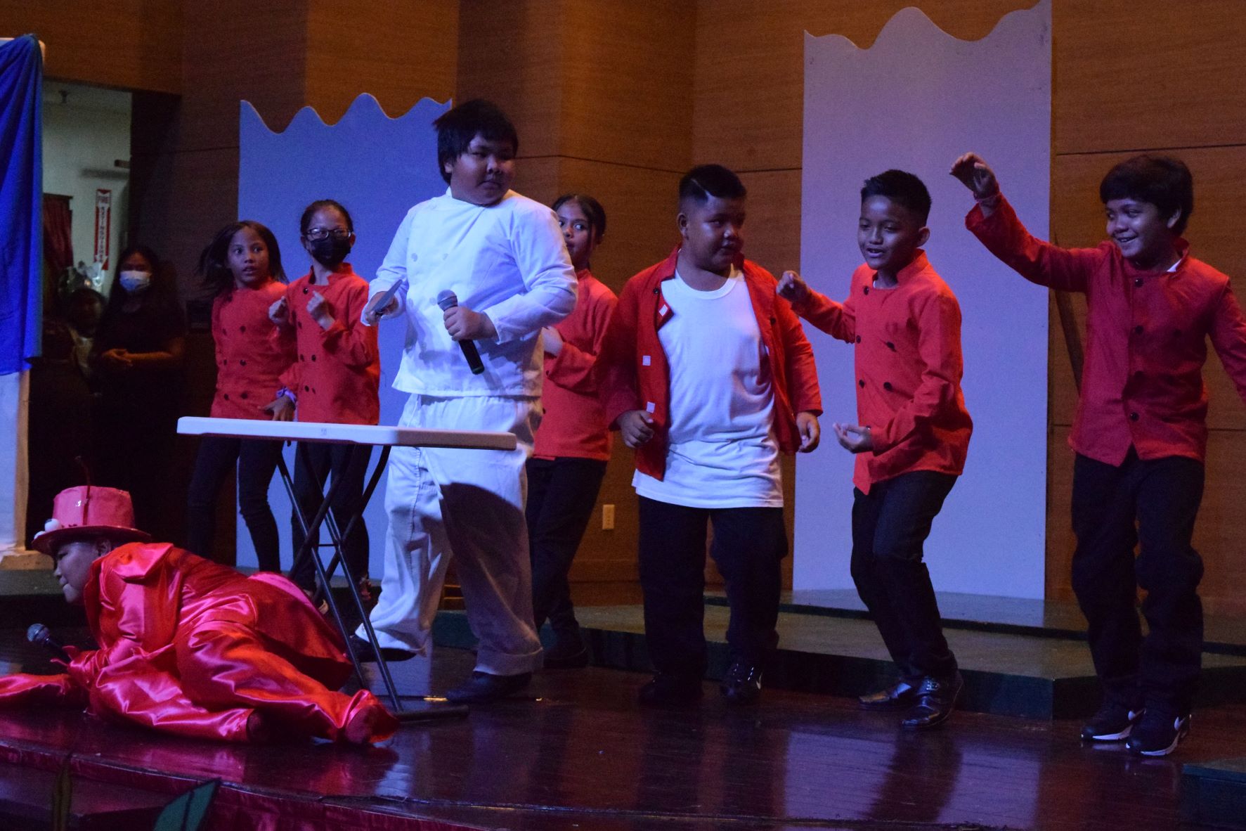 Sebastian lies on the floor as cooks Kianalise Sablan, Diane Merjilla, Nathan Coloma, Nicole Tangonan, Ridintor Coper, Lysander Domingo and Amlesh David Jha sing and dance in the Les Poissons scene.