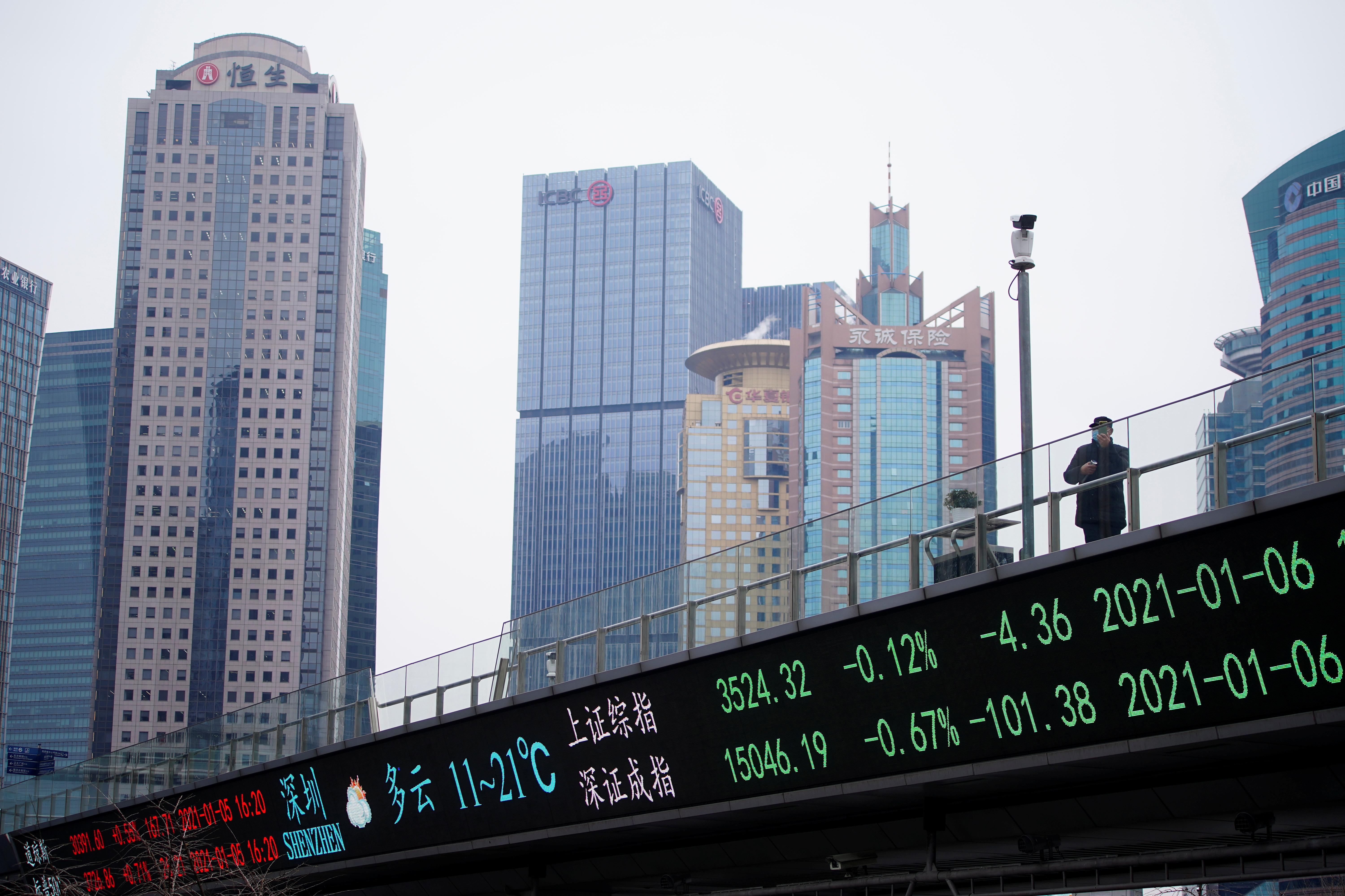 A man wearing a face mask stands on an overpass with an electronic board showing Shanghai and Shenzhen stock indexes, at the Lujiazui financial district in Shanghai, China, Jan. 6, 2021.