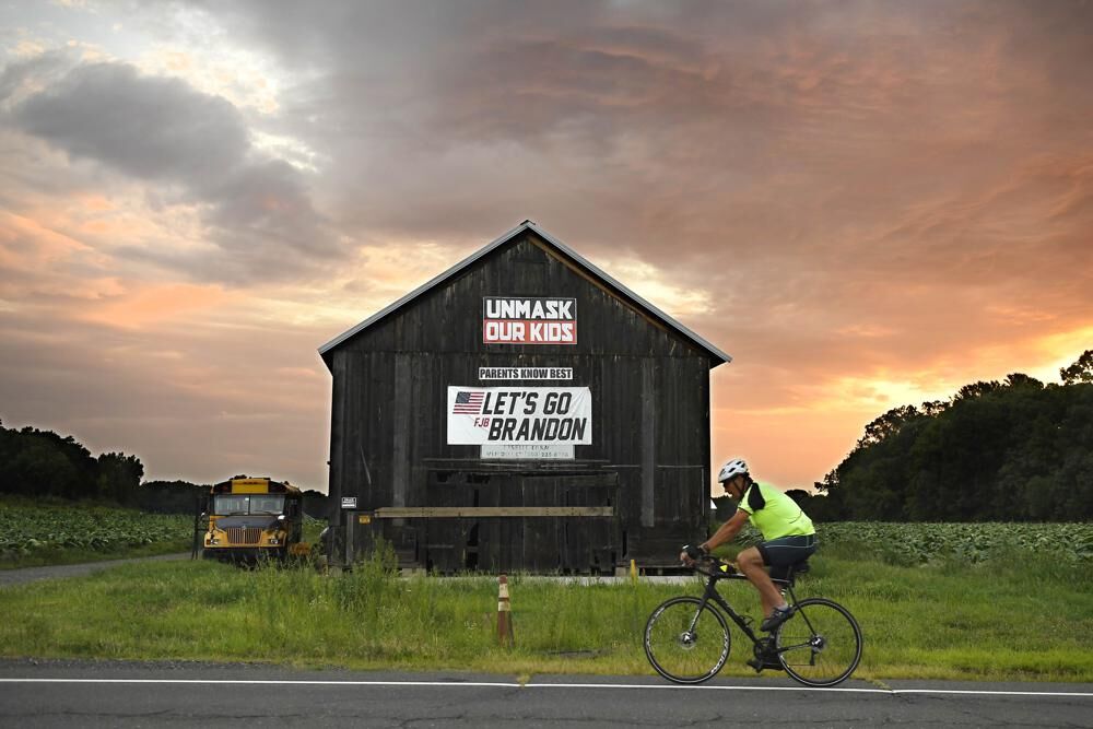 A bicyclist rides past a barn with political banner on it as the sun rises on primary election day, Tuesday, Aug. 9, 2022, in Suffield, Conn. Suffield is one of several small towns in Connecticut where control was flipped from Democrats to Republicans in 2021 municipal races.