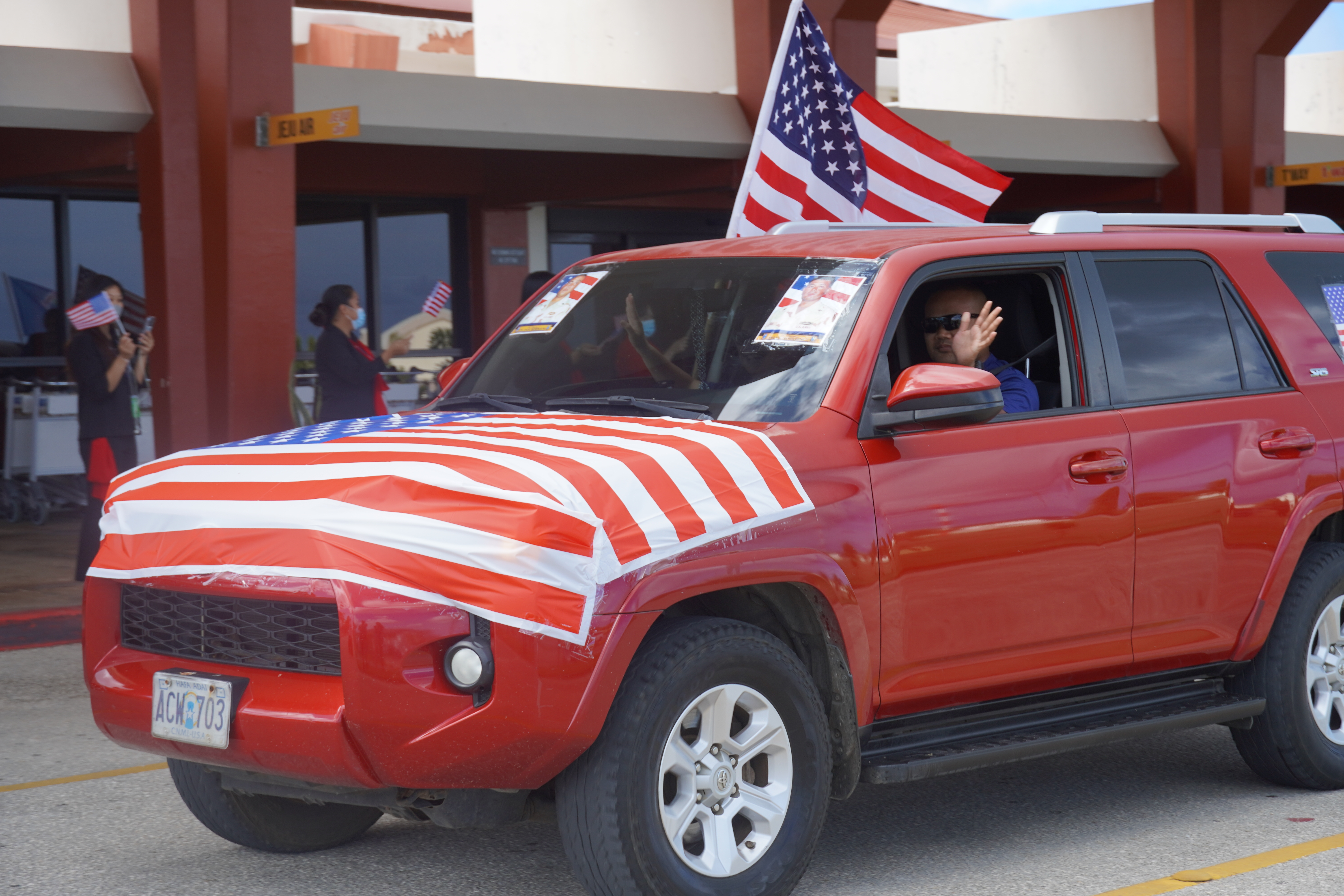 A motorcade participant waves from the driver's seat of his vehicle, adorned with the U.S. flag and photos of a fallen veteran.