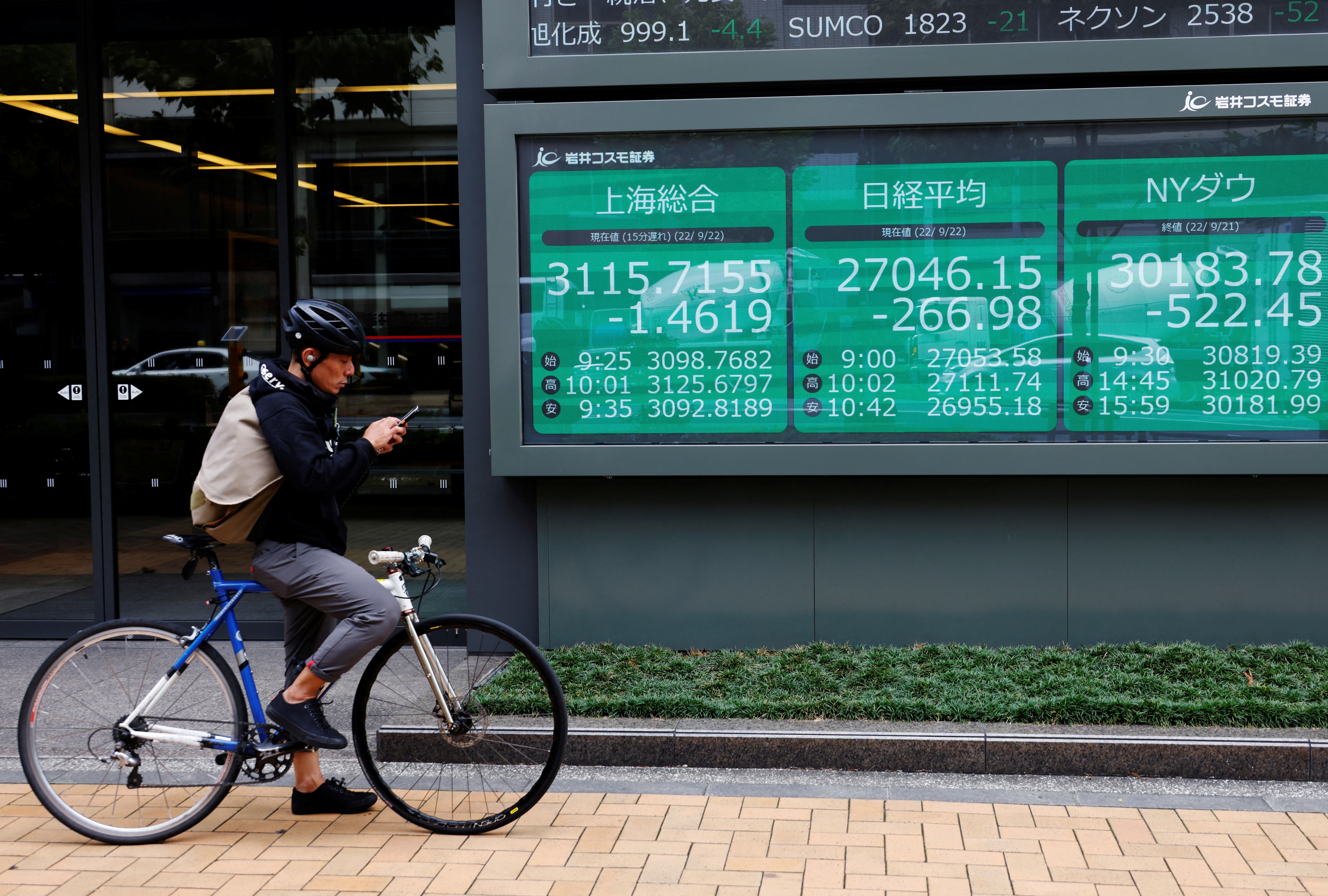 A man on a bicycle stands in front of an electronic board showing Shanghai stock index, Nikkei share price index and Dow Jones Industrial Average outside a brokerage in Tokyo, Japan, Sept. 22, 2022.