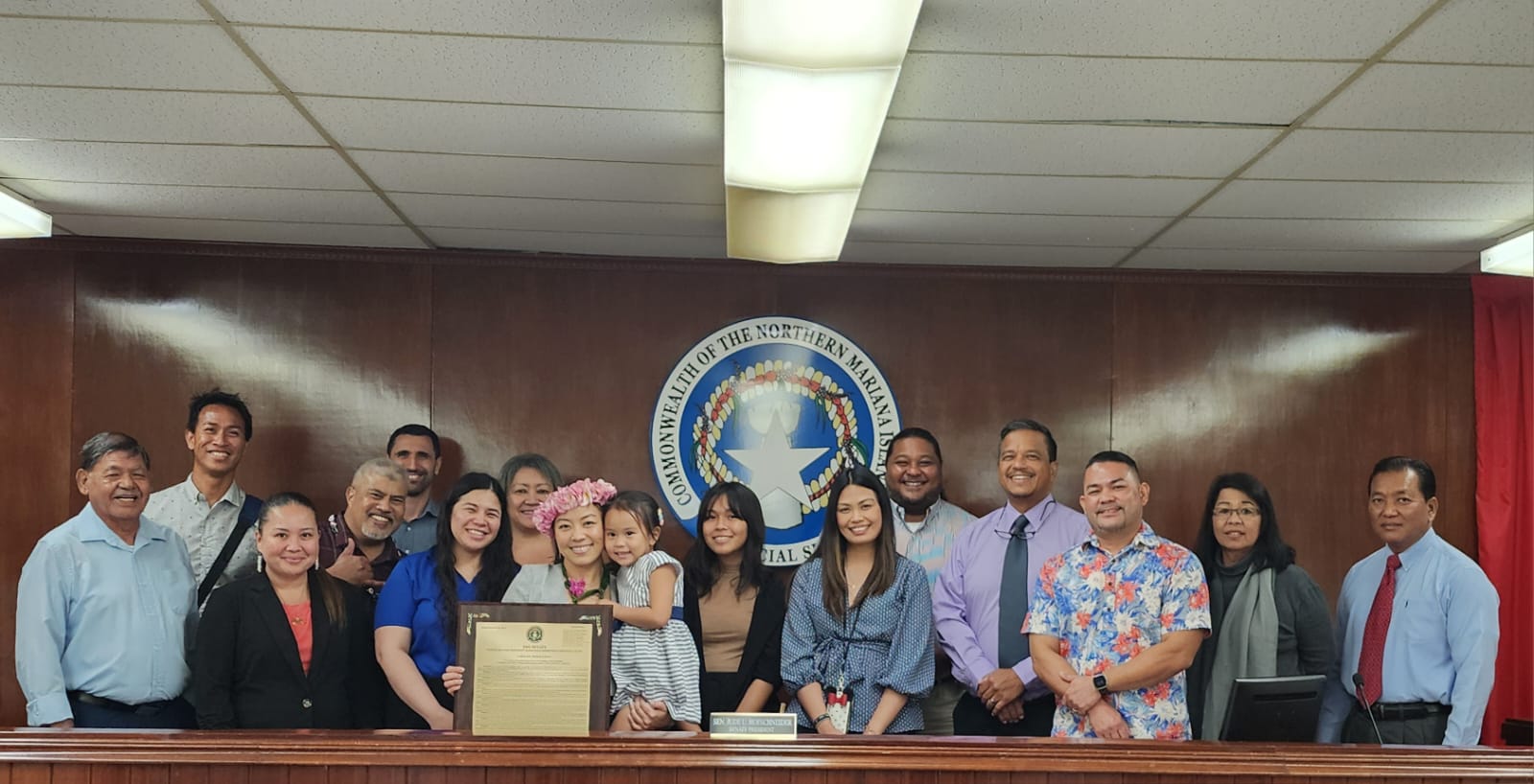 Hyun Jae Lee, wearing a mwar, holds a framed copy of a commemorative resolution adopted by the Senate to honor her exemplary service and selfless dedication to the Law Revision Commission and the NMI judiciary. With her in the photo are family members, friends and senators.