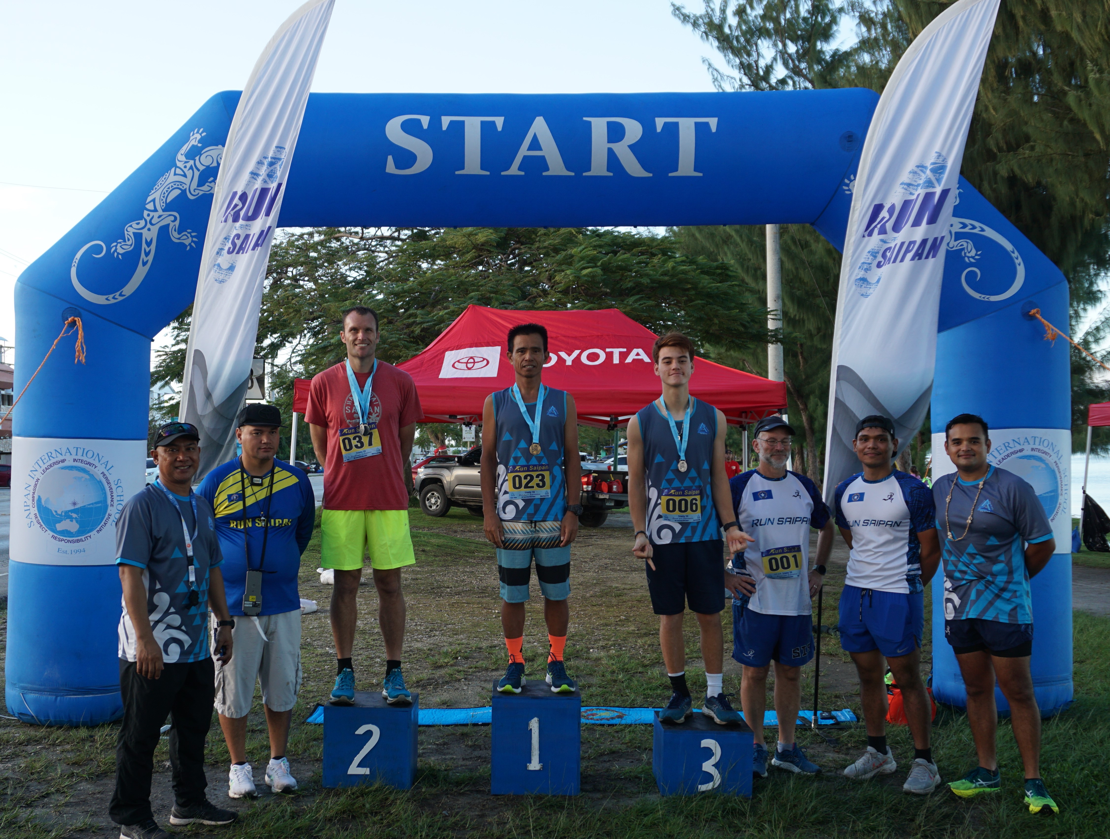 Joel Buco, Chad Taflinger and Theo Rodgers pose with their Day 2 medals and Run Saipan officials during the awards ceremony of the Run Saipan Trinity event Monday on  Beach Road in Oleai.