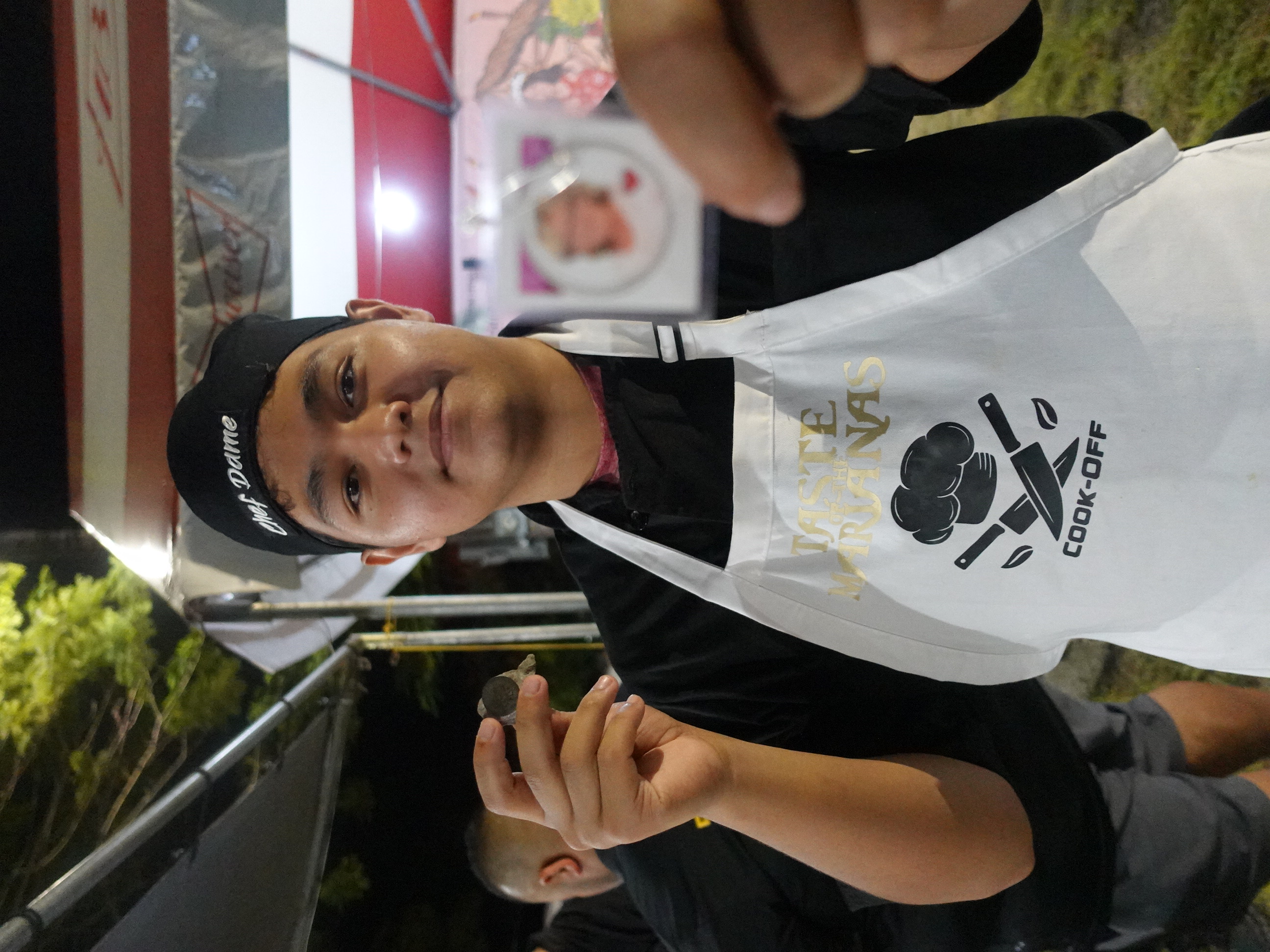 Damian Palacios, 1st place winner in the Hotel Association of the Northern Mariana Islands/NMTech Chamorro Bisteak Cookoff on June 24, 2023, displays a photo of his late grandmother and a lucky Irish rock from her that he carried in his pocket on his way to winning his first chefs competition.  The cookoff was held as part of the 24th Annual Taste of the Marianas International Food Festival & Beer Garden.