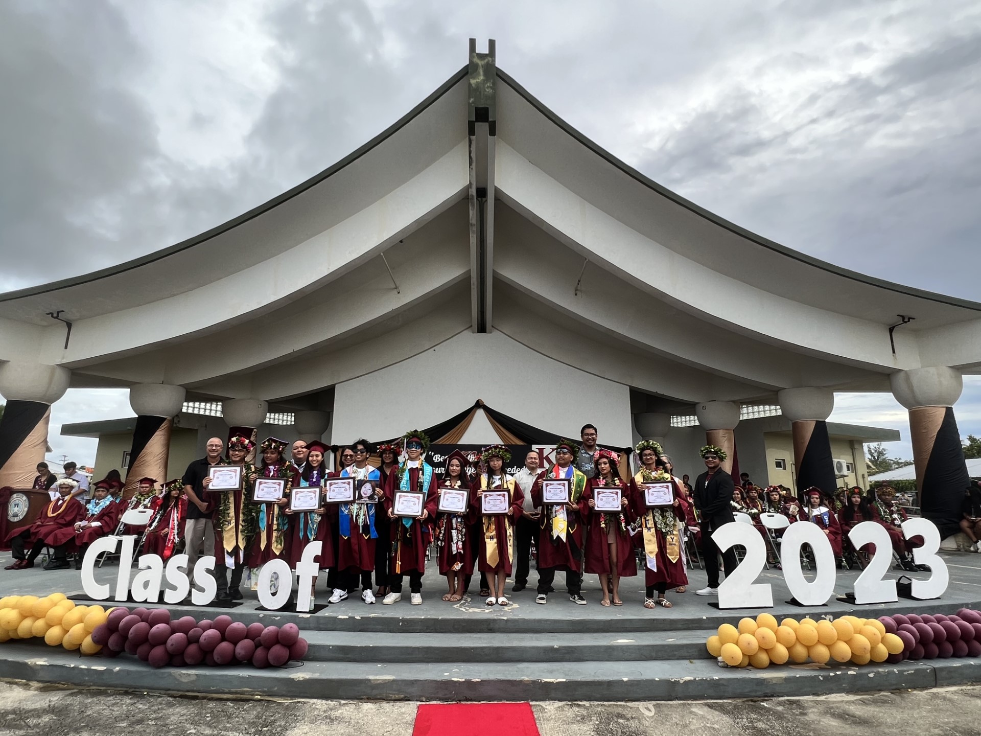 Kagman High School’s top 10 graduates: (1) Valedictorian Richard Carl Dela Cruz Lacson, (2) Salutatorian Kiyokazu Ma'gas Suzuki Gonzales, (3) Nanaka Joyeria Watanabe, (4) Jude Dominic Litulumar (5) D'anahlei Vifika Magofna Rodriguez (6) Jessica Nicole Tellei Pineda, (7) Xinyi Ni, (8) Zyron Altoveros Espiritu Santo, (9) Elicia Bernadette Mendiola Cabrera, and (10) Juneya Neiyongischigh Quitano. They are joined on the stage by Board of Education Chairman Antonio L. Borja, BOE Secretary/Treasurer Gregory Pat Borja, BOE Member Maisie B. Tenior, Teacher Representative Dr. Dora Borja Miura, Commissioner of Education Dr. Alfred B. Ada and Associate Commissioner for Administrative Services Eric Magofna.