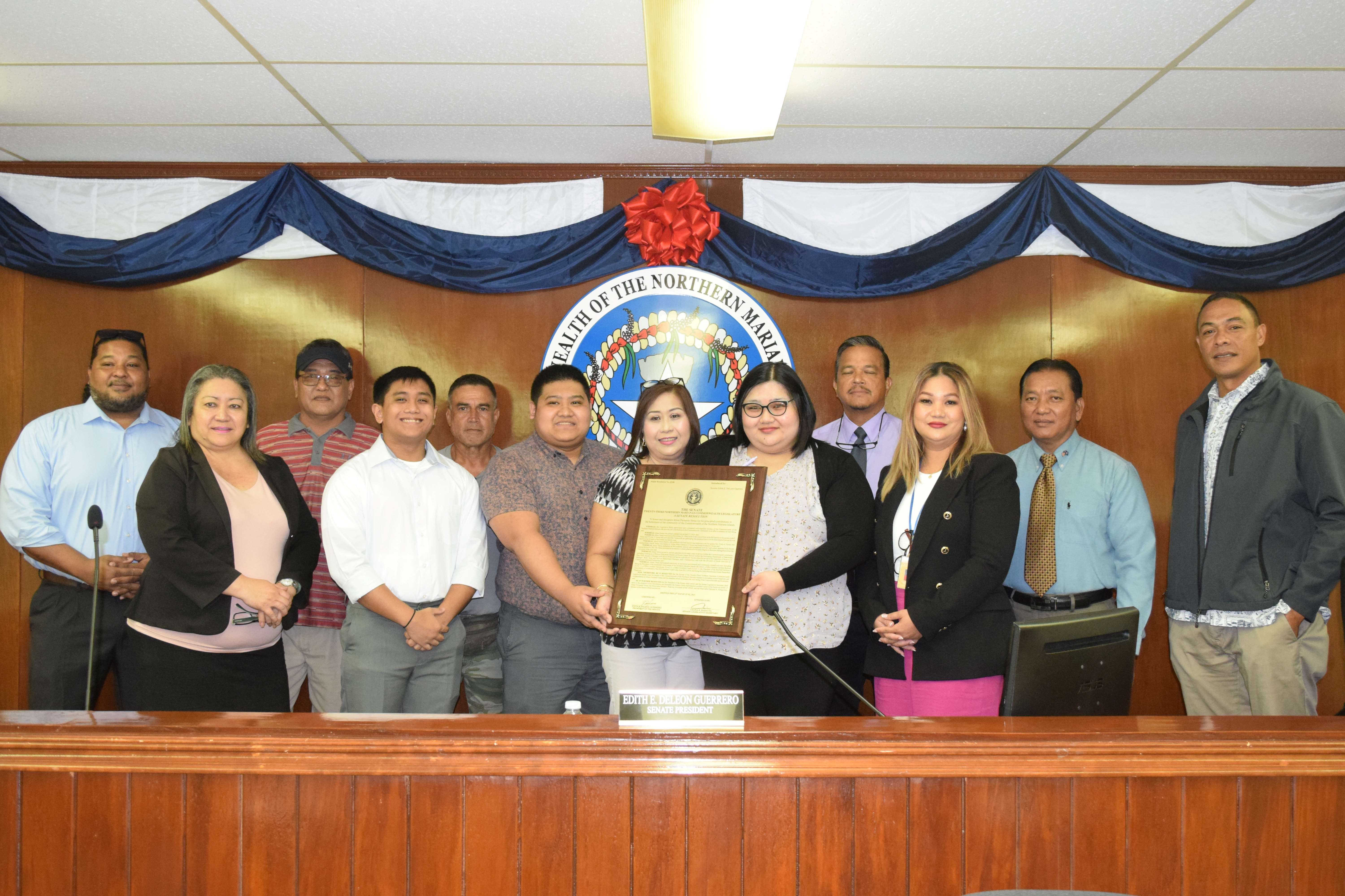 Cristy Venus, center, Marc Christian and Ruby Ann hold the framed copy of Senate Resolution 23-6 as they pose with senators for a photo in the Senate chamber on Friday. The resolution honors the late Basketball Association of NMI President Abner Fernando Venus.