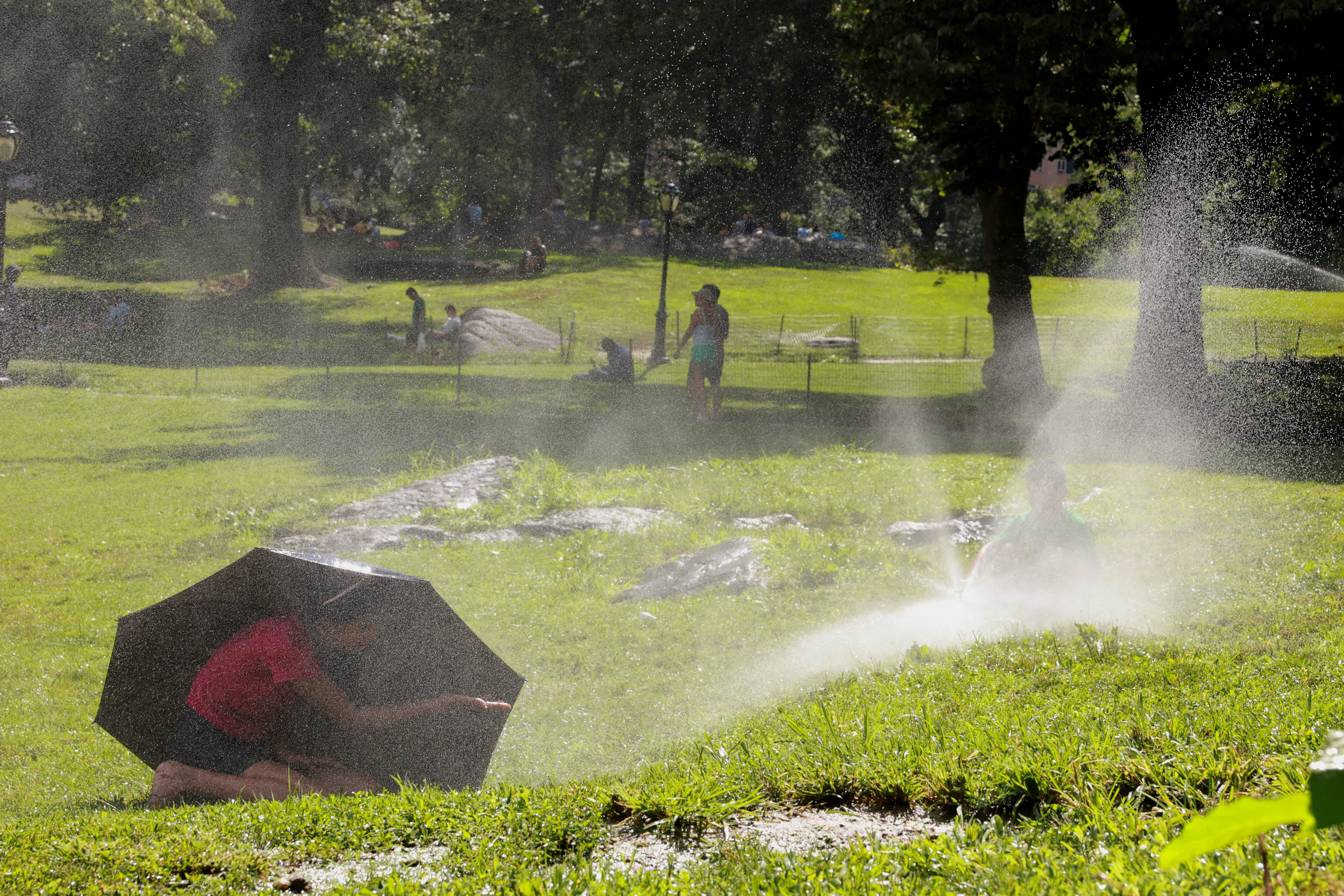 A child takes shelter under an umbrella while another one plays with a water sprinkler, in Manhattan's Central Park, in New York City, U.S., July 28, 2023. REUTERS/Amr Alfiky