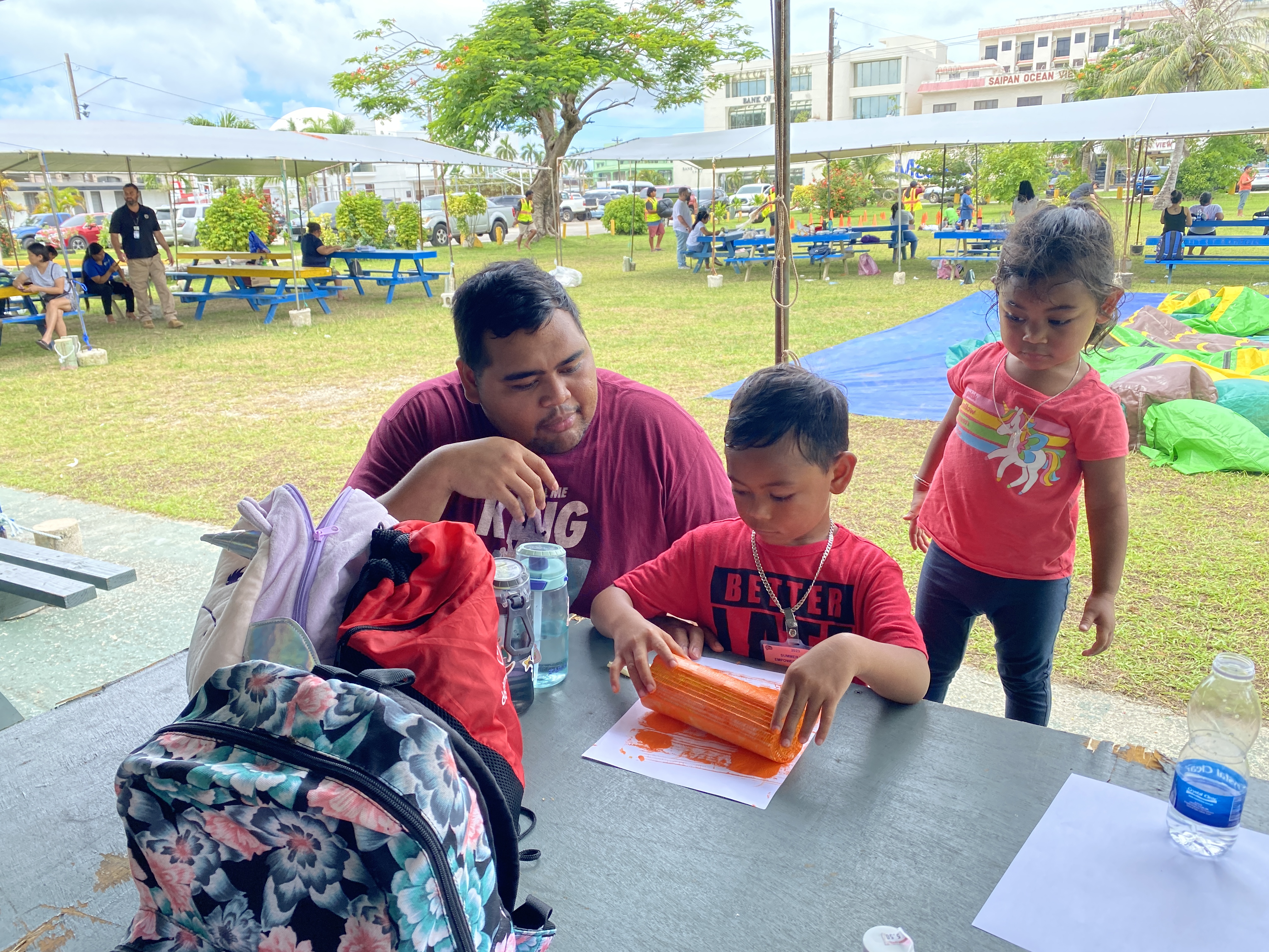 RJ Mendiola and his daughter Bryånna watch as her brother Bryce paints a picture using a banana stalk at the DYS 2023 Summer Youth Empowerment Camp