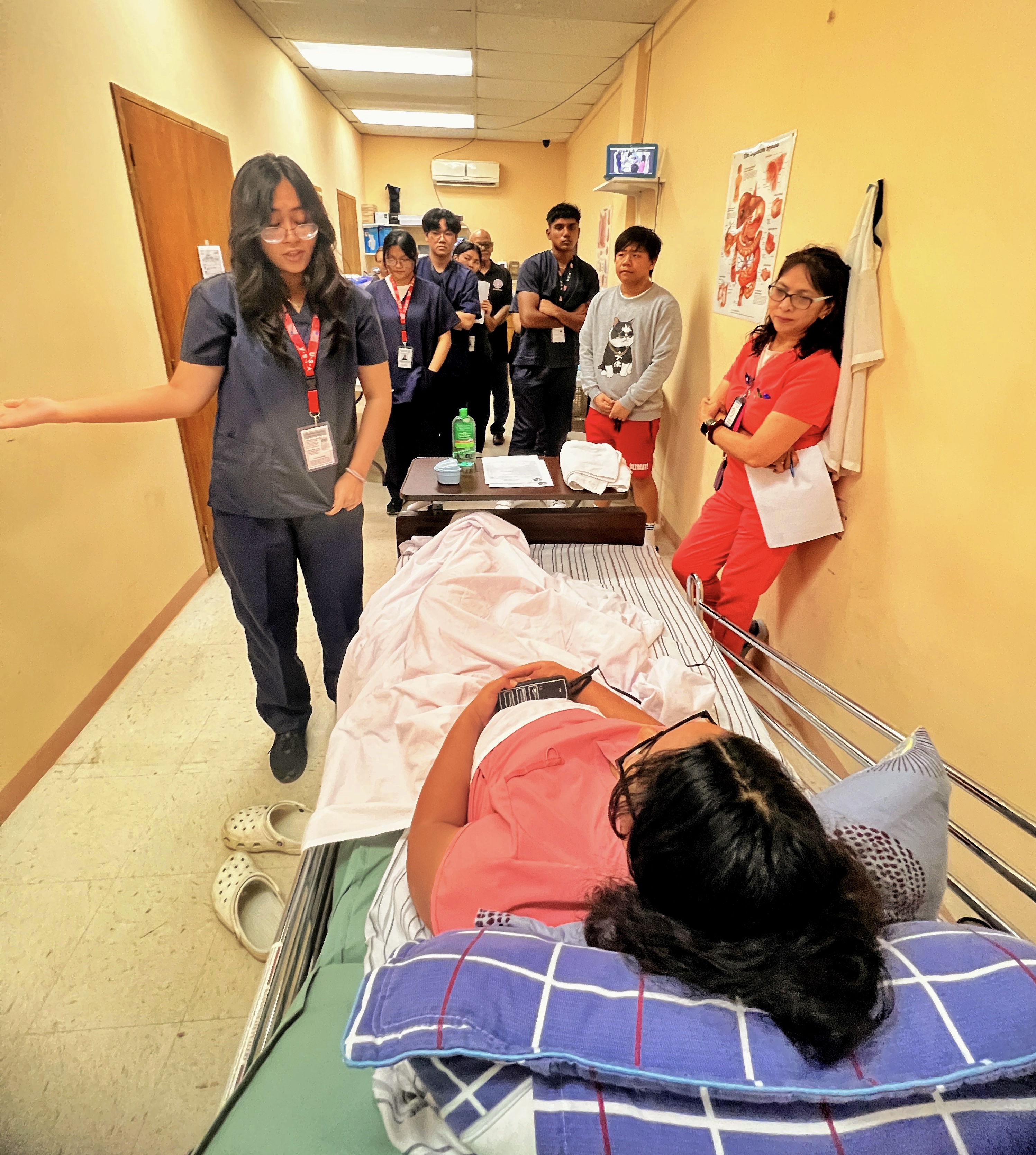 In the presence of their instructor, nursing professional Vivian Concepcion, standing right,  PSS-CTE Nursing Assistantship Program participant Elizah Canlas, left, gestures as she talks to her “patient,” program participant Freya Greathouse, while their classmates observe the hands-on training session.