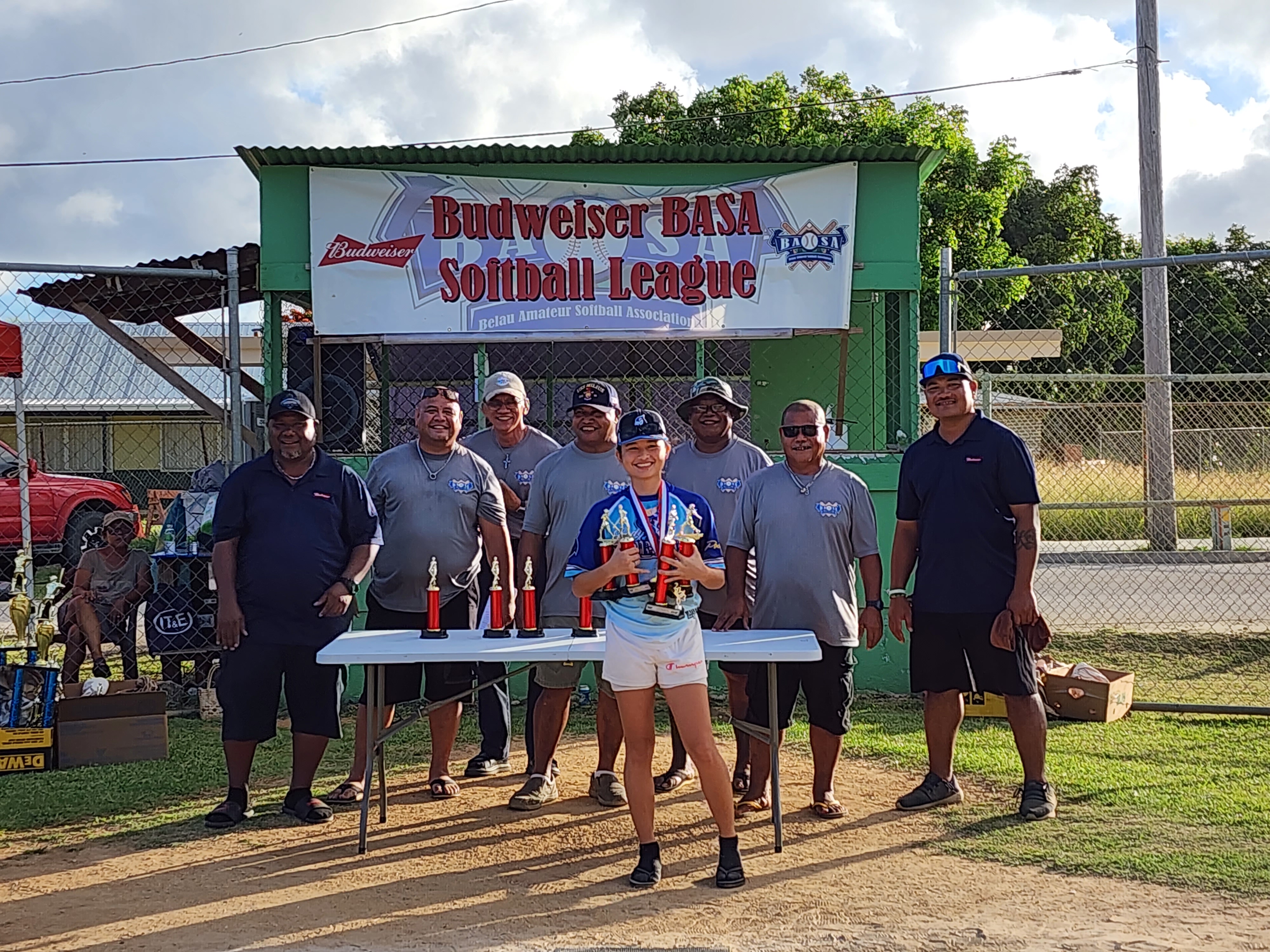Kiana "Baby K" Camacho holds four of her five individual trophies as she poses for a photo with the 2023 Budweiser Belau Amateur Softball Association Softball League officials during the awards ceremony  at the Dandan baseball field on Sunday.