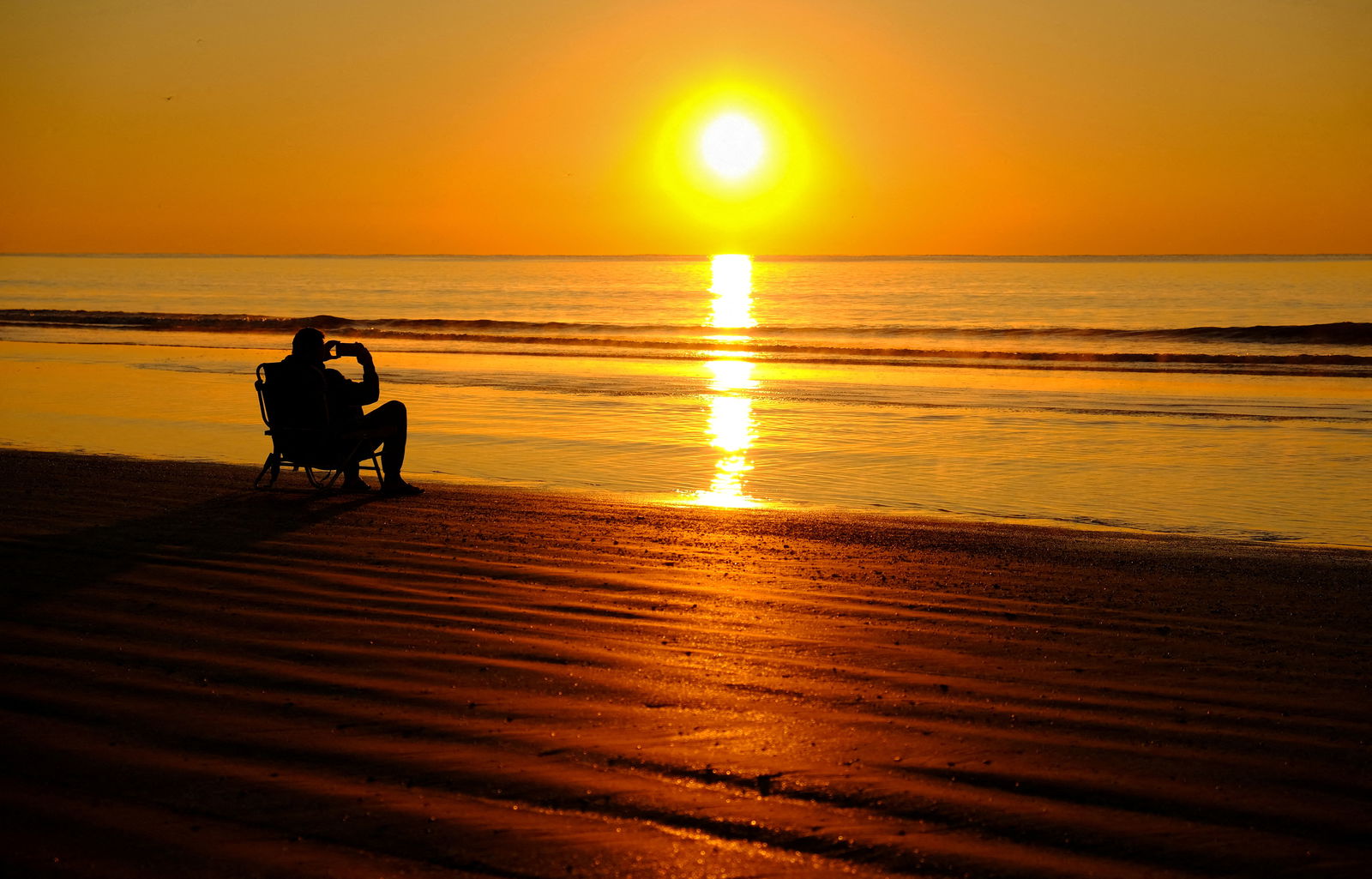 Sitting on a beach at water's edge, a man takes a photo as the sun rises over Hilton Head Island, South Carolina, U.S., March 16, 2023. 