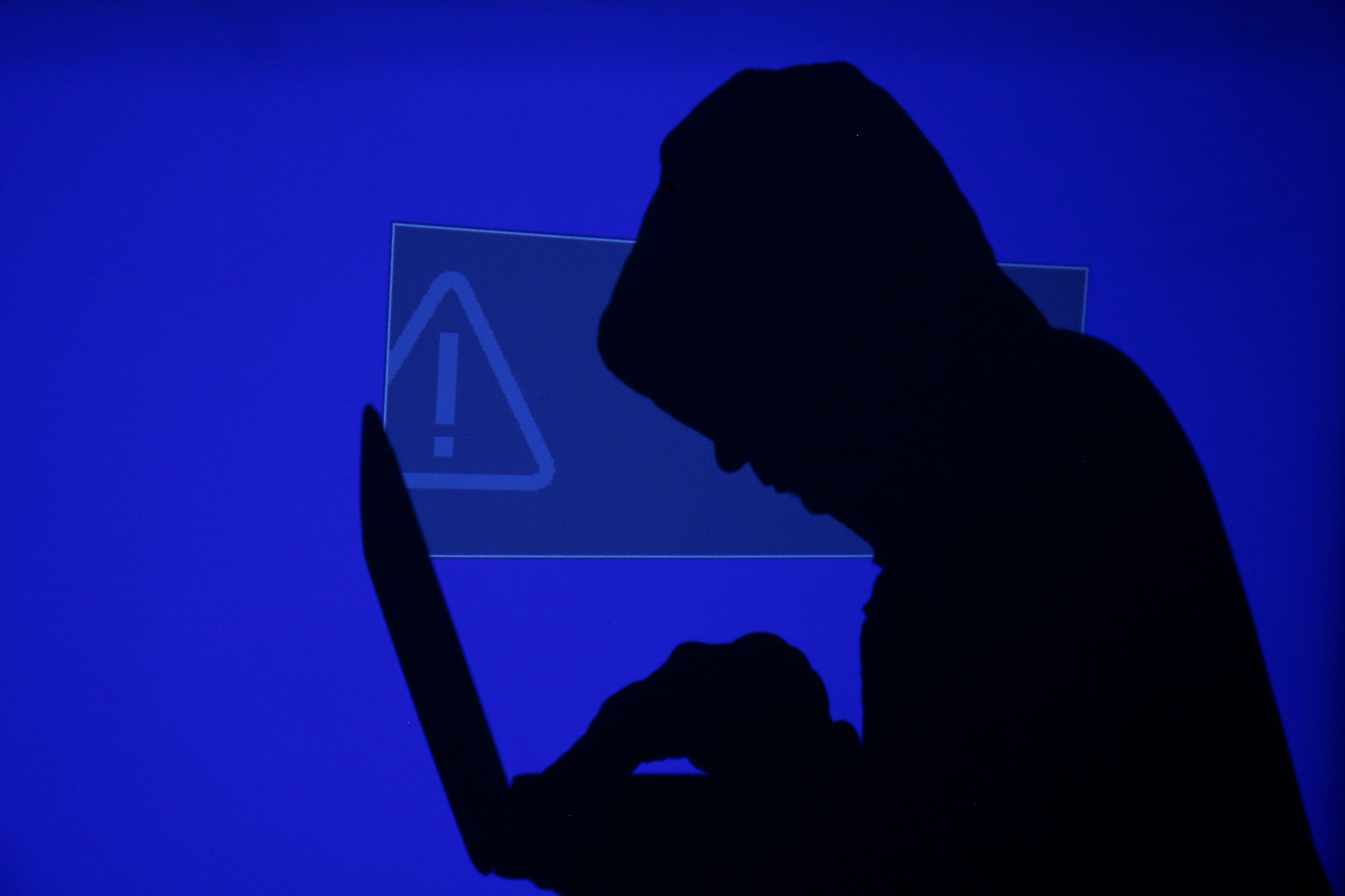 A hooded man holds a laptop computer as blue screen with an exclamation mark is projected on him in this illustration picture taken on May 13, 2017. Capitalizing on spying tools believed to have been developed by the U.S. National Security Agency, hackers staged a cyber assault with a self-spreading malware that has infected tens of thousands of computers in nearly 100 countries. 