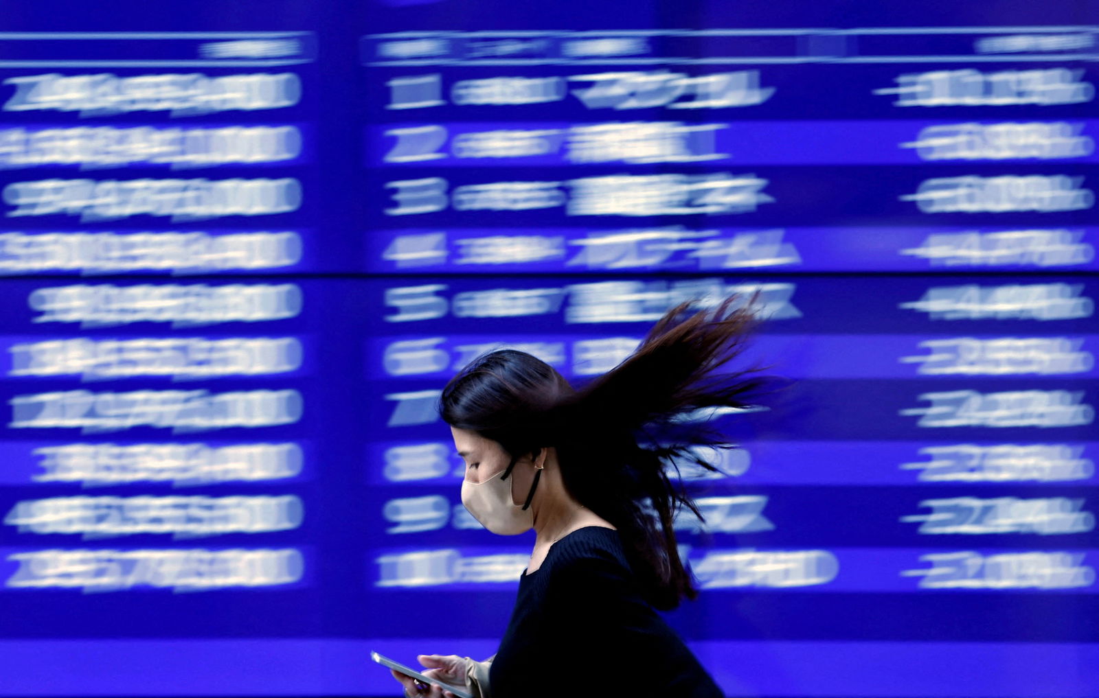 A passerby walks past an electric monitor displaying recent movements of various stock prices outside a bank in Tokyo, Japan, March 22, 2023.