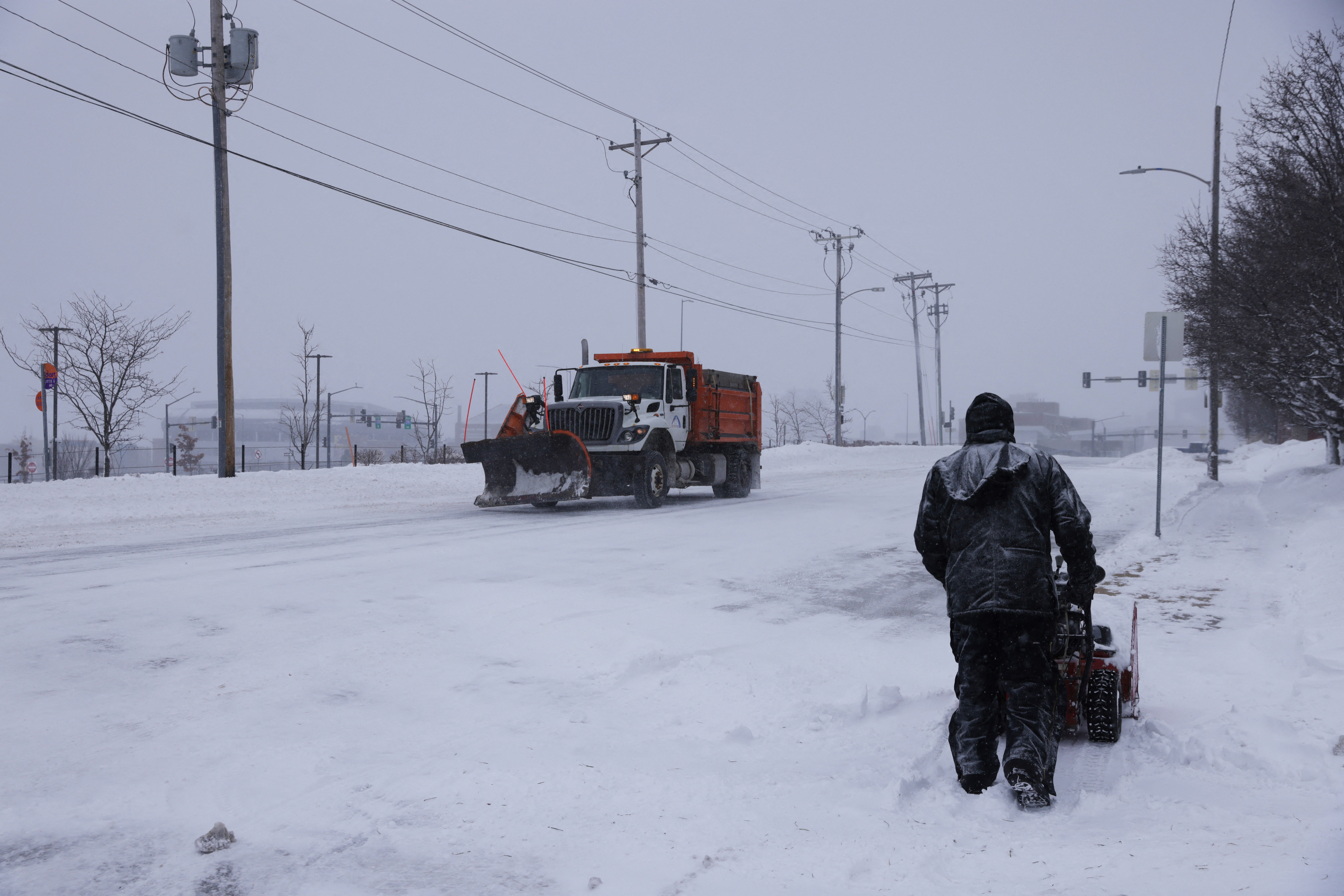 A worker removes snow from from a sidewalk as a plow passes by after a blizzard left several inches of snow in Des Moines, Iowa, U.S., January 13, 2024. 