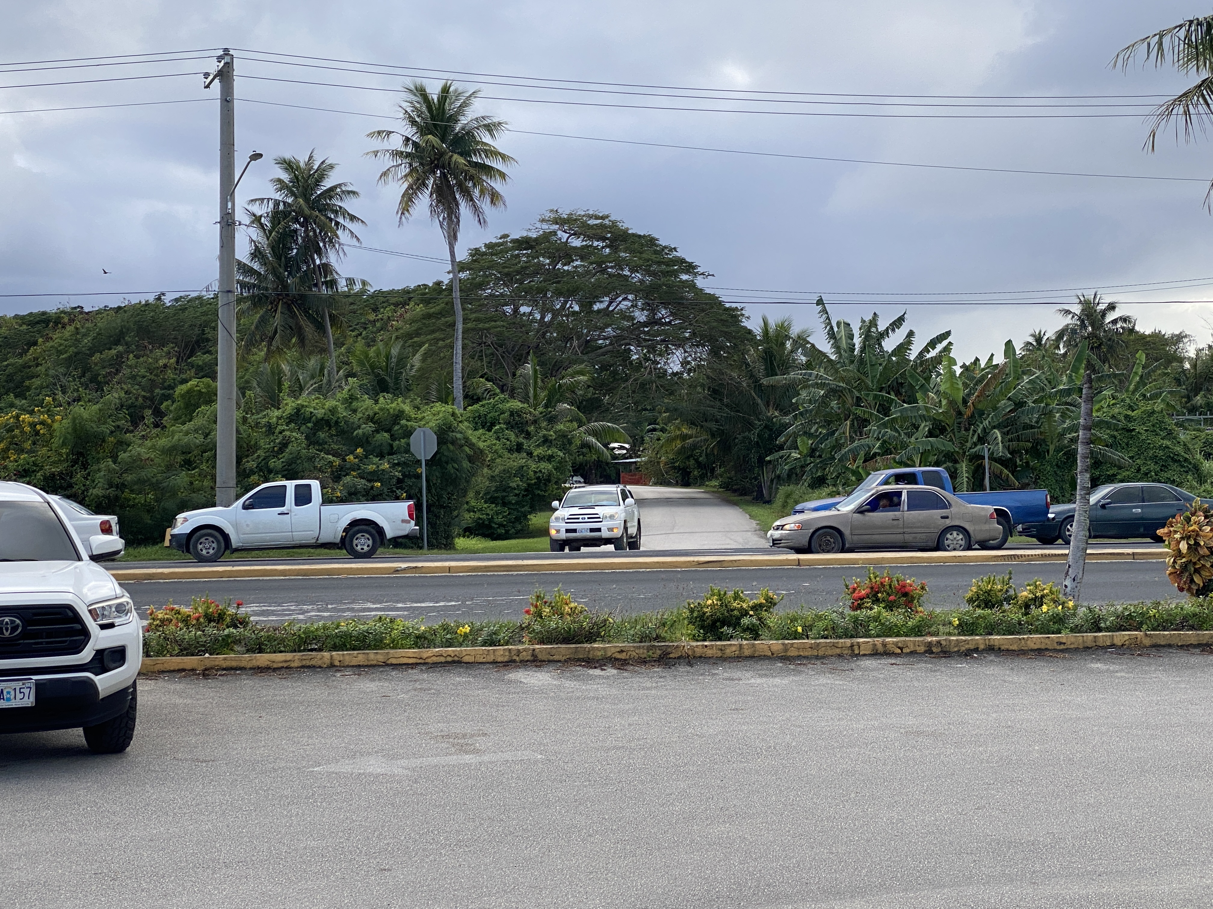 A vehicle exits Texas Road to make its way to Chalan's Kiya three-way intersection. Behind the vehicle, Texas Road stretches past homes and Oleai Elementary School before it connects to a secondary road. 