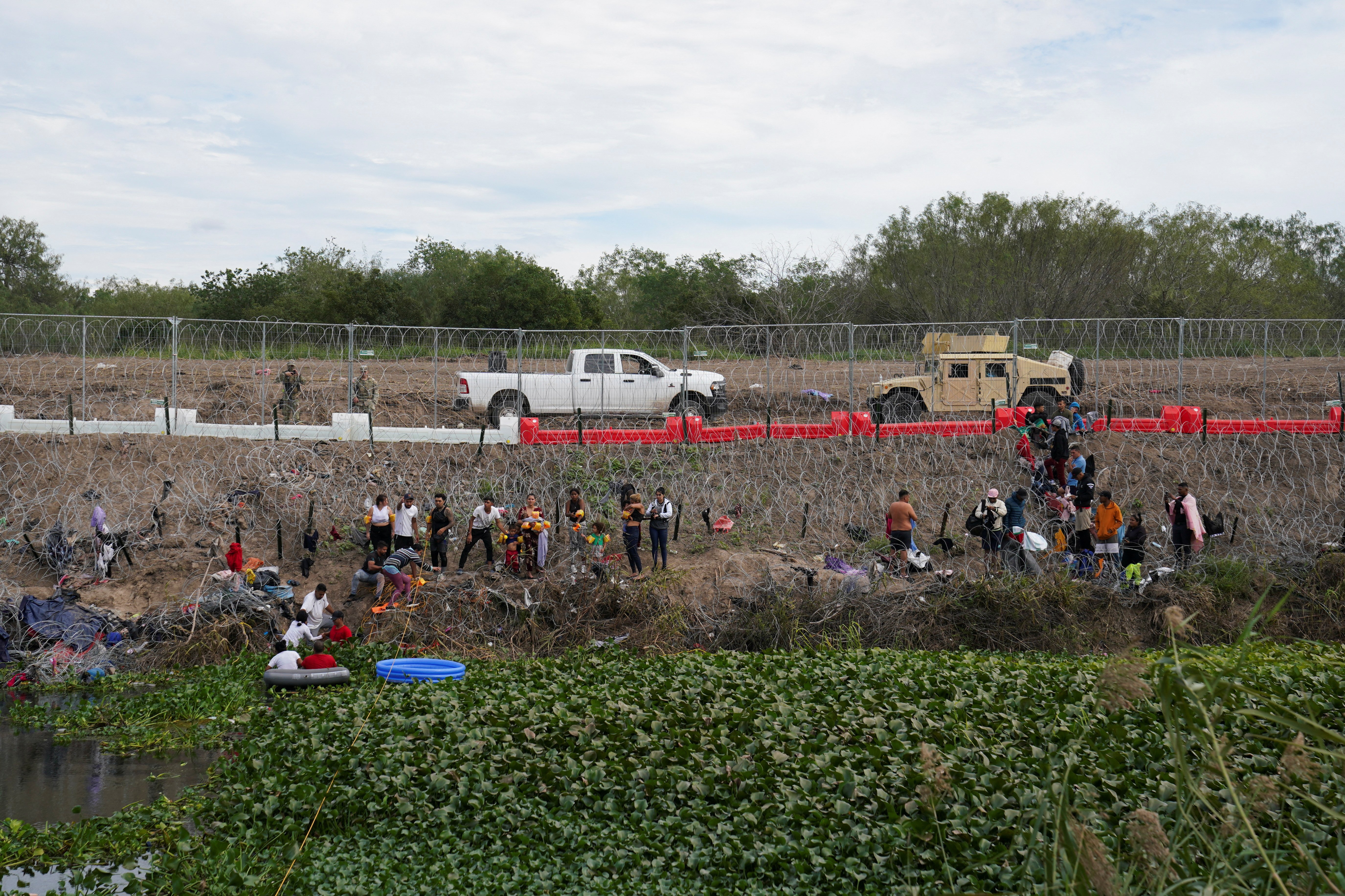 People wait behind concertina wire and newly installed barriers to turn themselves to the U.S. border authorities, in Brownsville, Texas, as seen from Matamoros, Mexico, December 29, 2023. 
