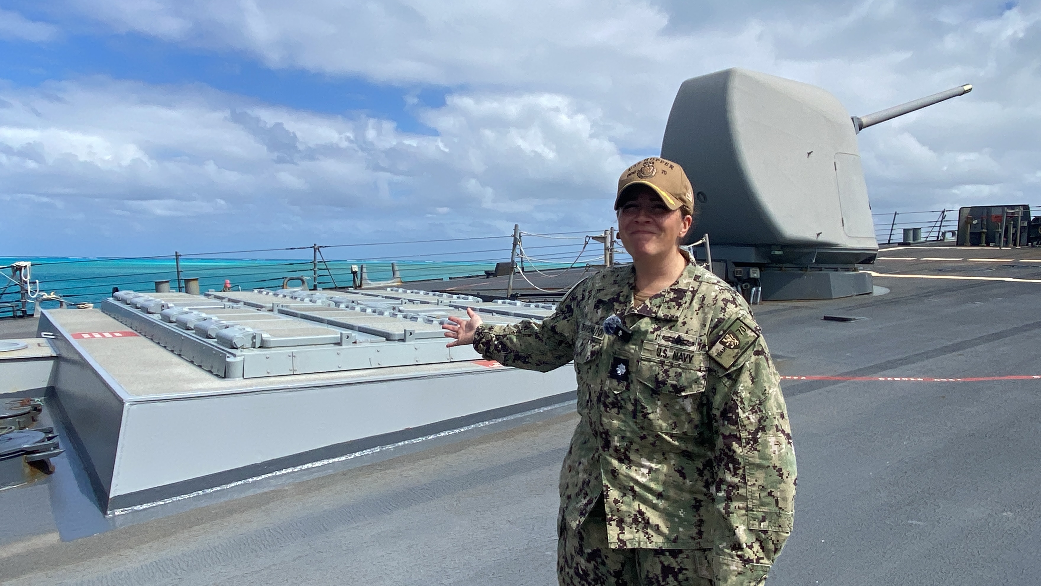 CDR Andrea Benvenuto gestures toward the vertical launch system cells that carry various missiles aboard the USS Hopper. Behind her is a five-inch gun.