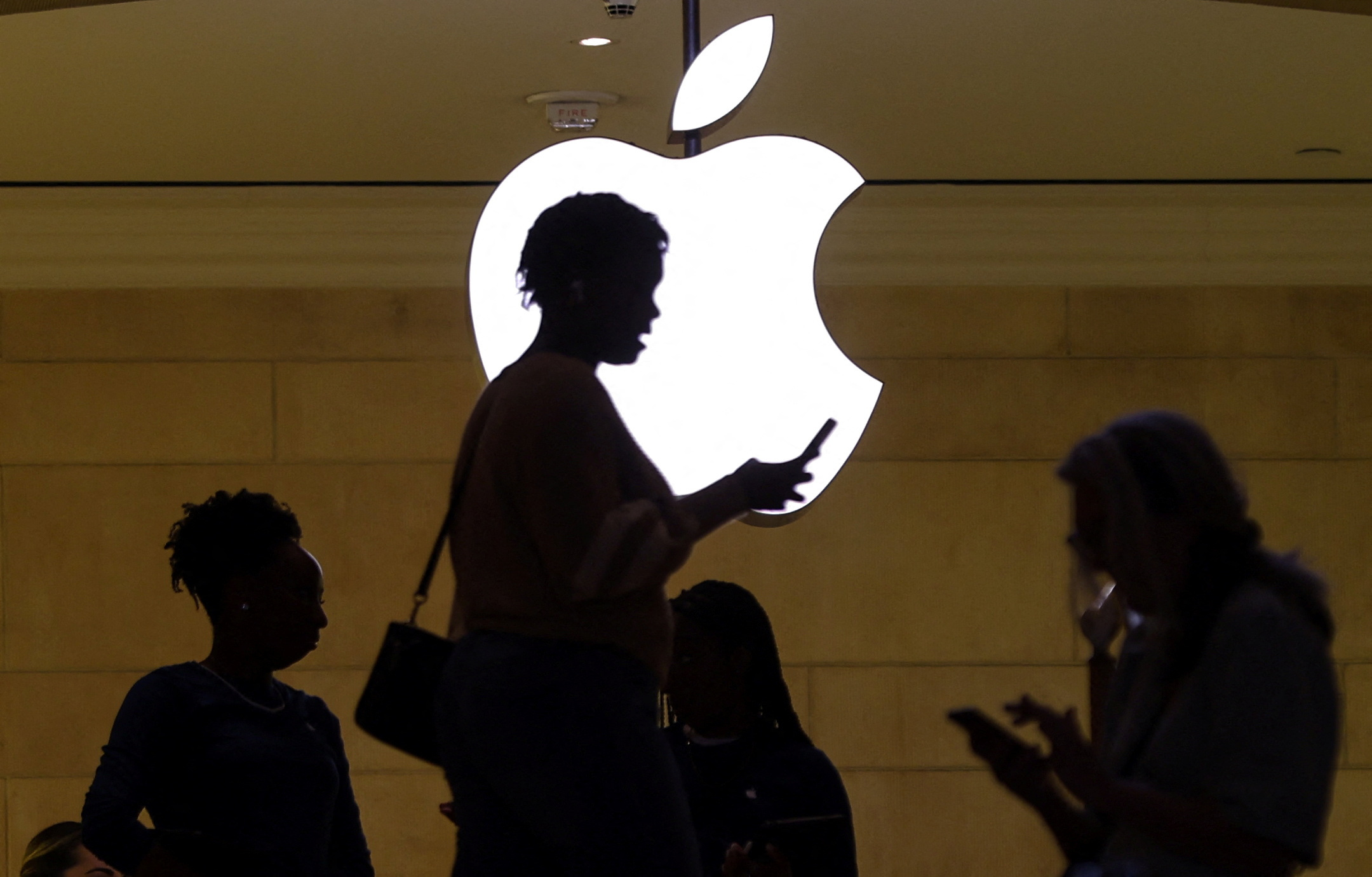 Women uses an iPhone mobile device as she passes a lighted Apple logo at the Apple store at Grand Central Terminal in New York City, U.S., April 14, 2023. 