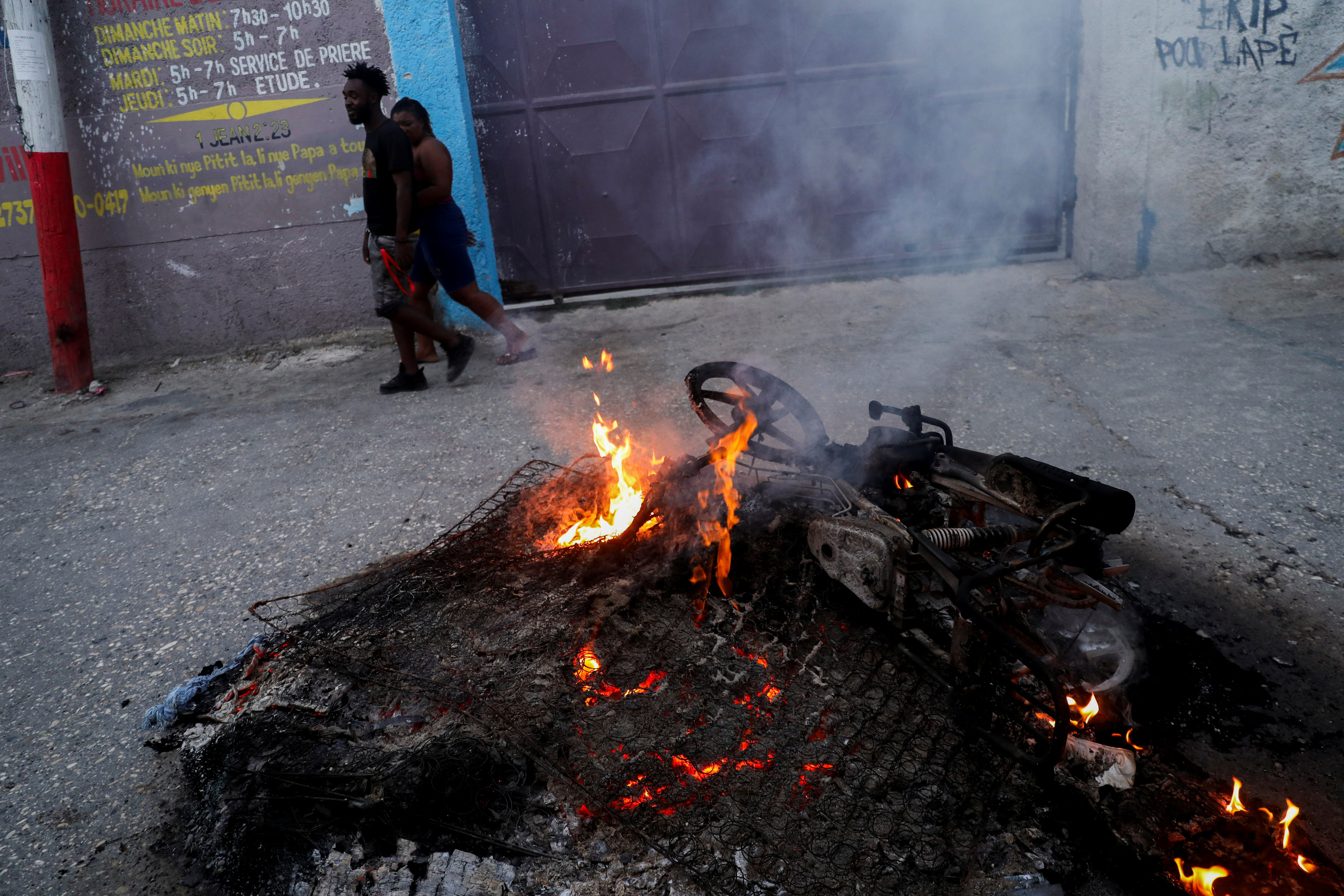 People walk past a pile of burning mattresses and the remains of the motorcycle of suspected gang leader Makandal who was killed and set on fire by local residents, amid an escalation in gang violence, in Port-au-Prince, Haiti March 20, 2024. 