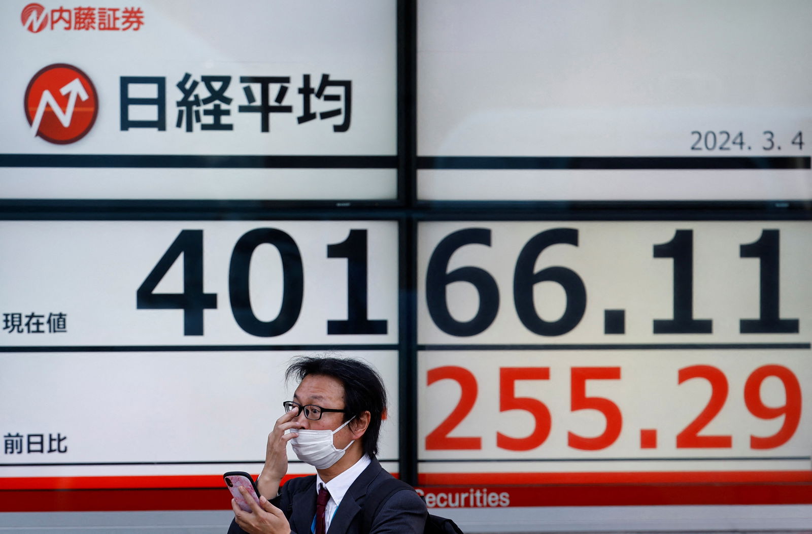 FILE PHOTO: A man uses a smartphone in front of an electronic screen displaying Japan's Nikkei share average outside a brokerage in Tokyo, Japan March 4, 2024. 