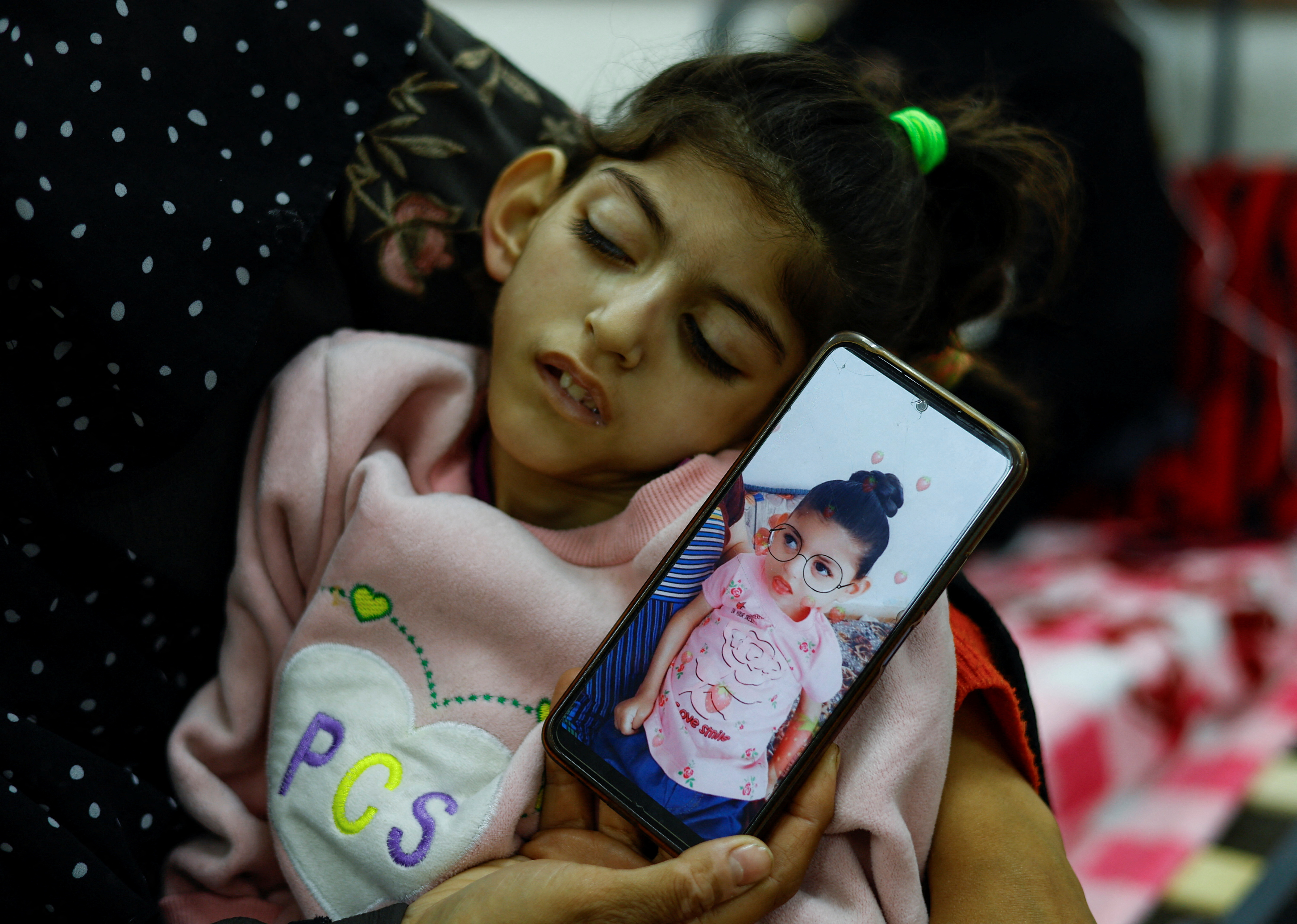 Palestinian woman Umm Mesbah Heji holds her malnourished daughter Israa, who is quadriplegic and epileptic, as she displays on a smartphone a picture showing Israa before the ongoing Israel-Hamas conflict, at al-Awda health center in Rafah in the southern Gaza Strip, March 12, 2024. 