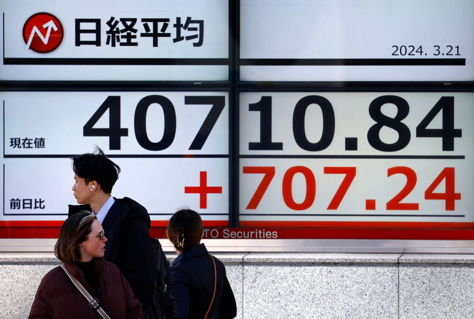 Passersby walk in front of an electric screen displaying Japan's Nikkei share average outside a brokerage in Tokyo, Japan on March 21, 2024.