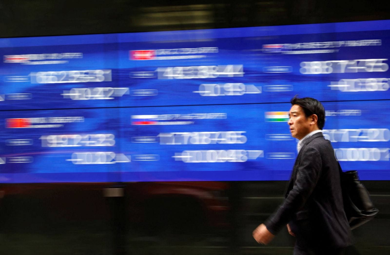 A passerby walks past an electric monitor displaying various countries’ stock price indices outside a bank in Tokyo, Japan, March 22, 2023.