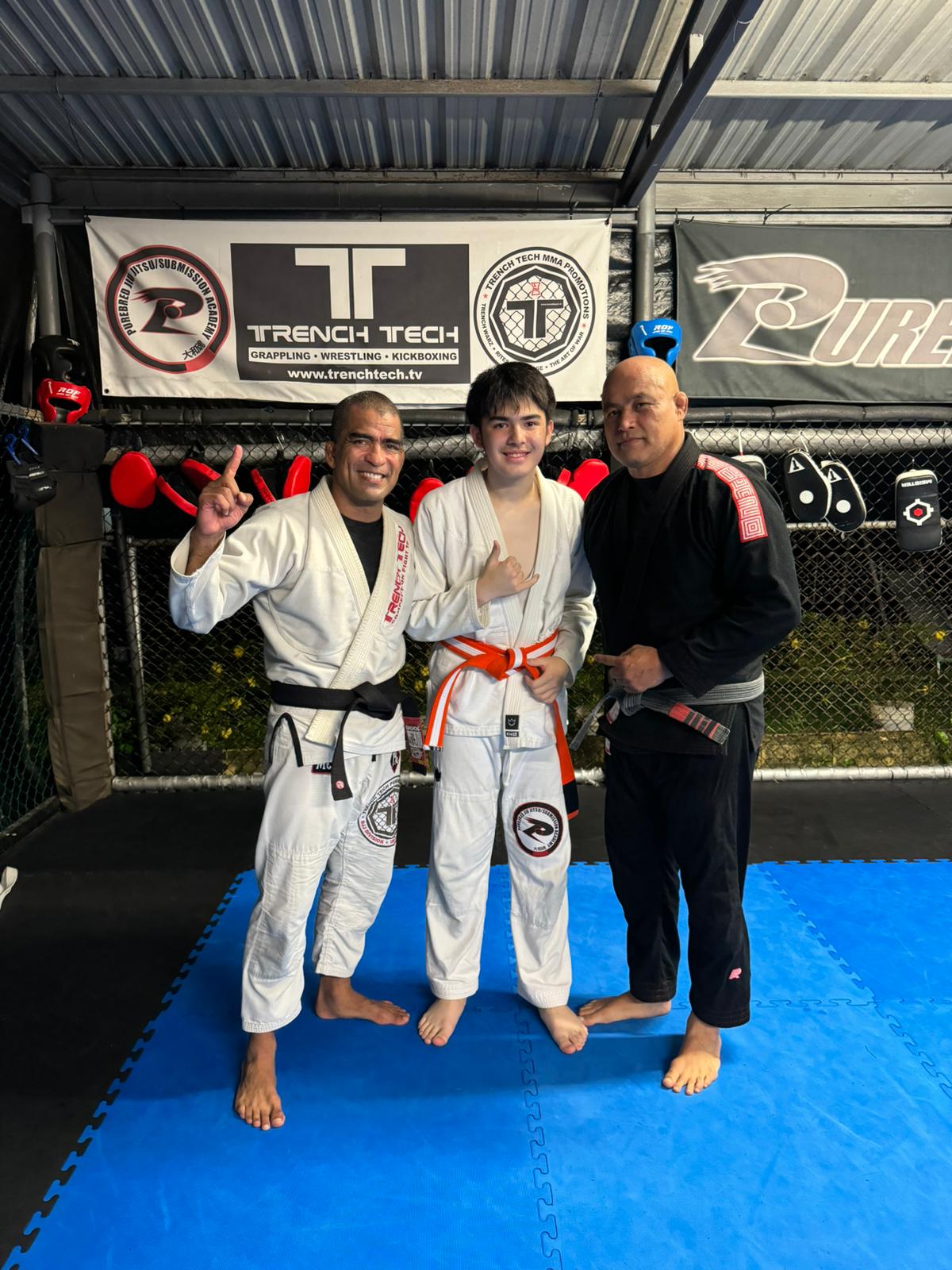 Gabriel Mitchell, center, poses with his newly earned juvenile white/orange belt alongside instructors Cuki Alvarez and Steven Roberto during a BJJ class at the Trench Tech gym.
