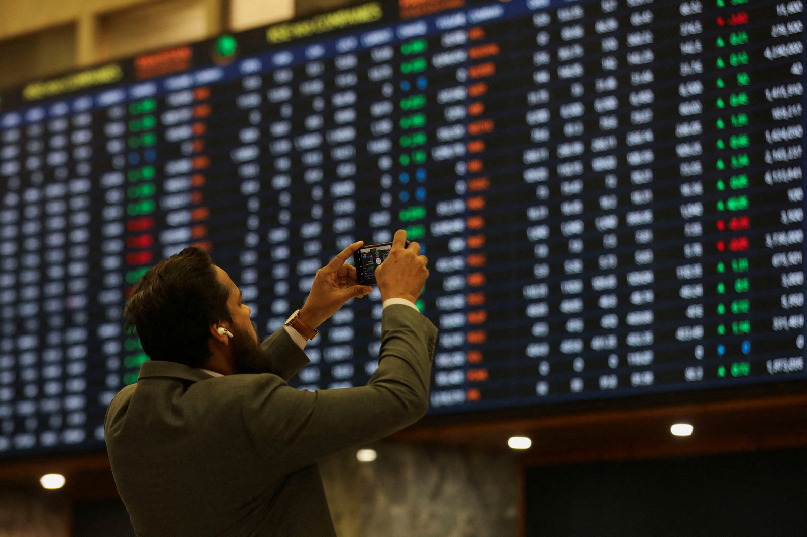 A man uses a mobile phone as he takes a photo of the electronic board displaying share prices during a trading session at the Pakistan Stock Exchange, in Karachi, Pakistan on Nov. 28, 2023.