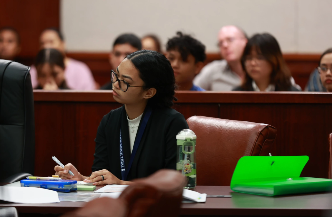 Saipan Southern High School competitor McKelly Reyes prepares notes during the opposing team’s examination of a witness during Preliminary Round 1, supported by her teammates and coaches Kassandra Knoff and Brendan Layde behind.