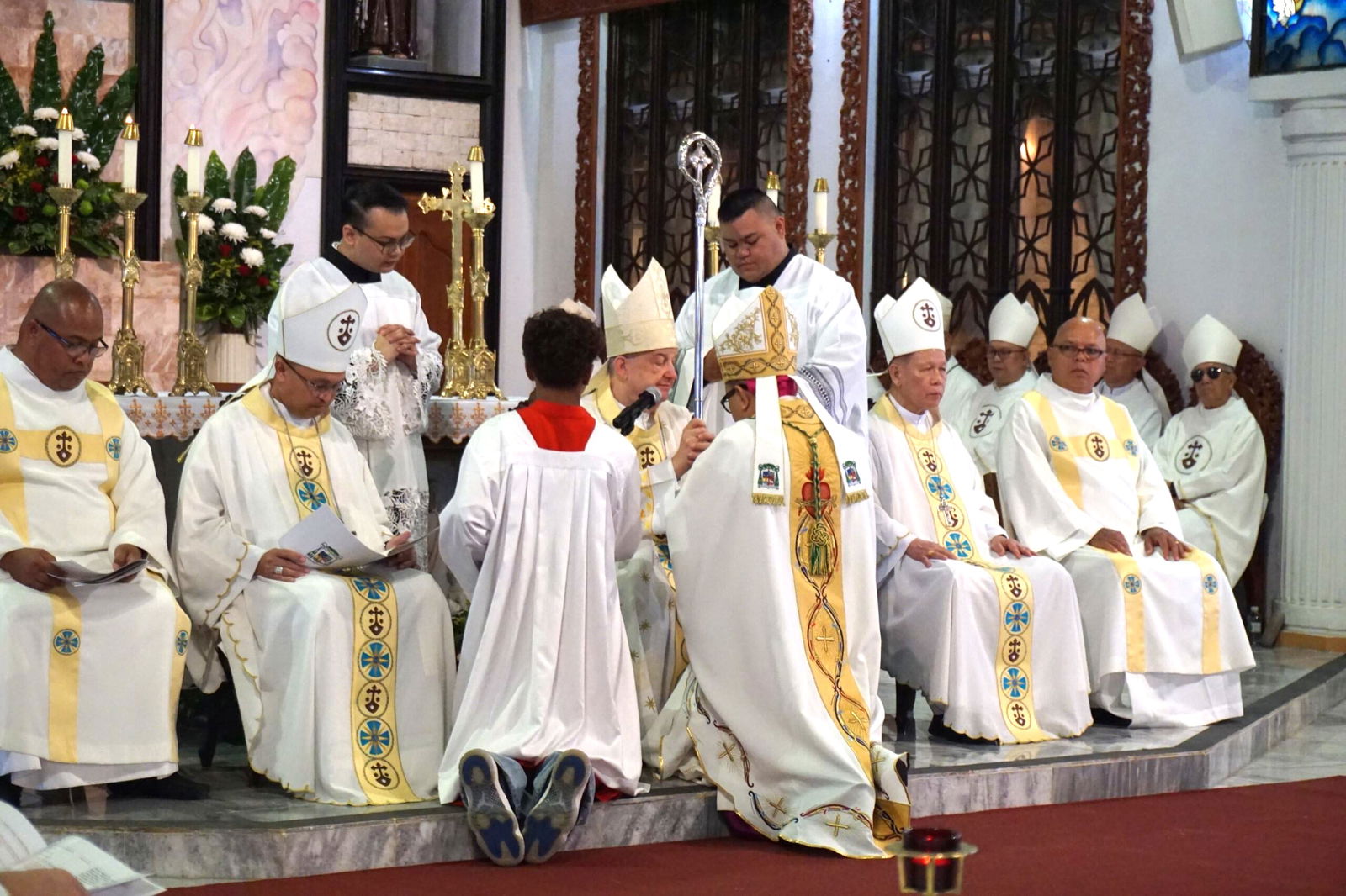 Bishop Romeo Convocar, kneeling, back to the camera, receives his bishop’s pastoral staff.