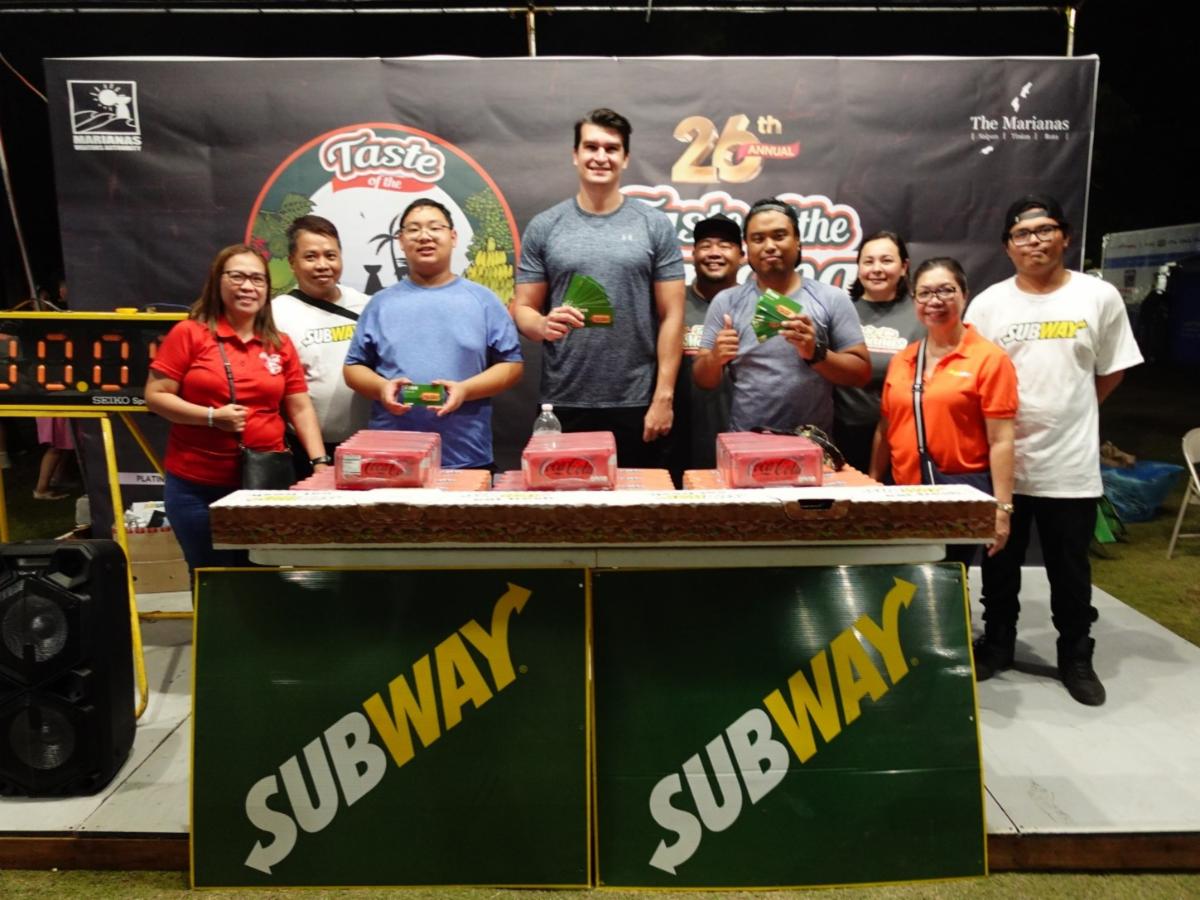 Jeremiah Rother, center front, finished an 18-inch sub in less than 20 minutes to take 1st place in the Subway Sandwich Challenge at the 26th Annual Taste of the Marianas International Food Festival on May 3, 2025, in Saipan. Also pictured are 2nd place finisher Eubo “Bob” Liu, third left; 3rd place finisher Simon Necesito, fourth right; and representatives of the Marianas Visitors Authority and event sponsor Subway/Coca-Cola/Foremost.