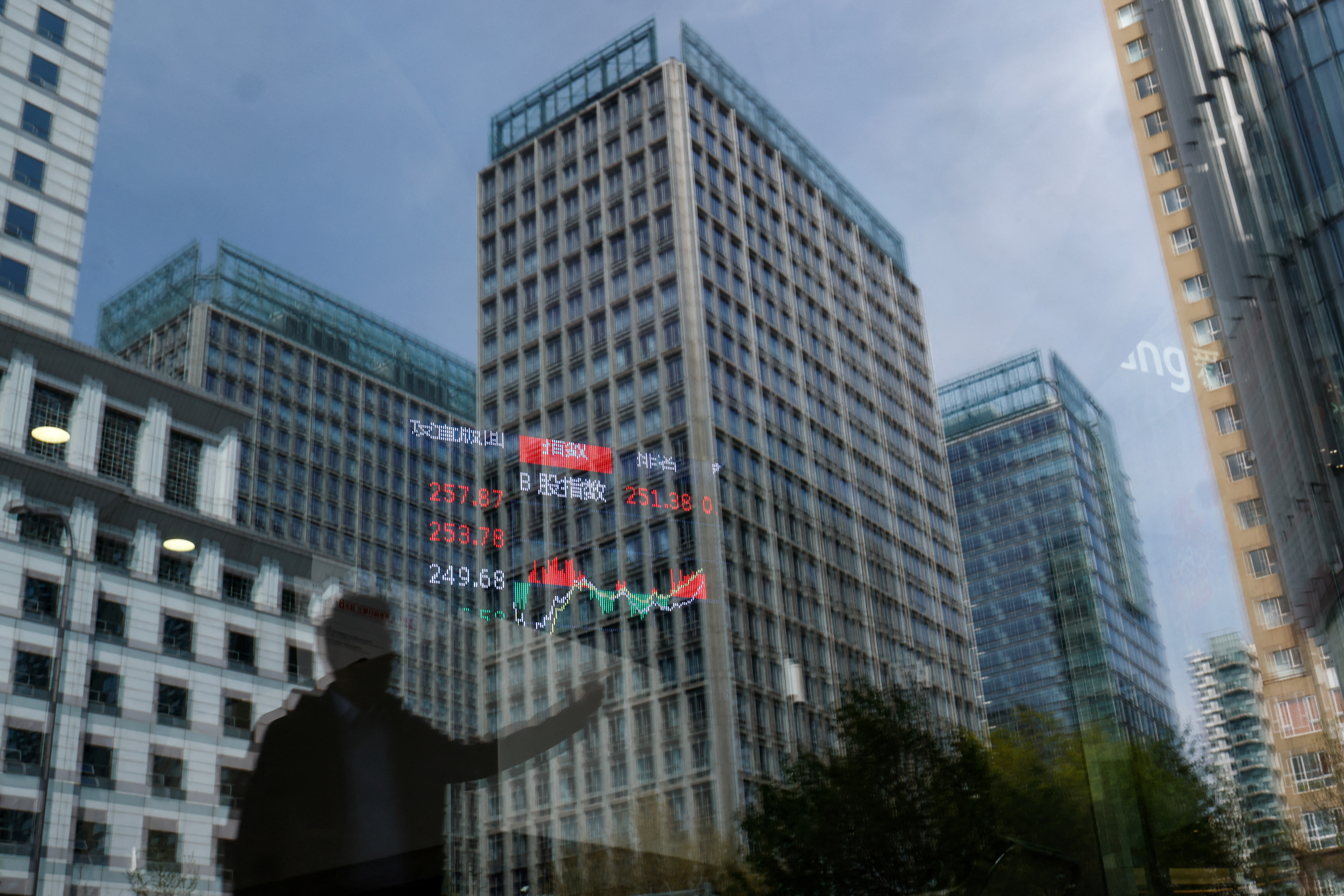 A person and buildings are reflected on the glass of a brokerage house where a display board is hung inside showing the stock index information, in Beijing, China on April 9, 2025.REUTERS