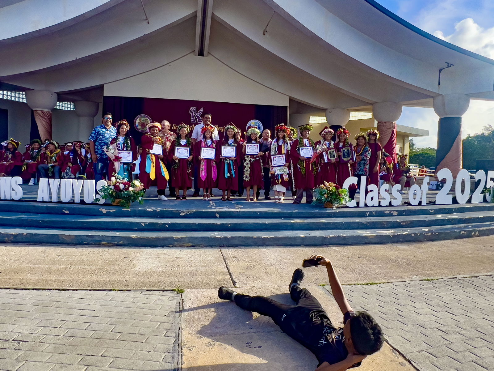 In the foreground, a family member of one of Kagman High School’s top 10 graduates from the Class of 2025 lies on the ground to take photos.