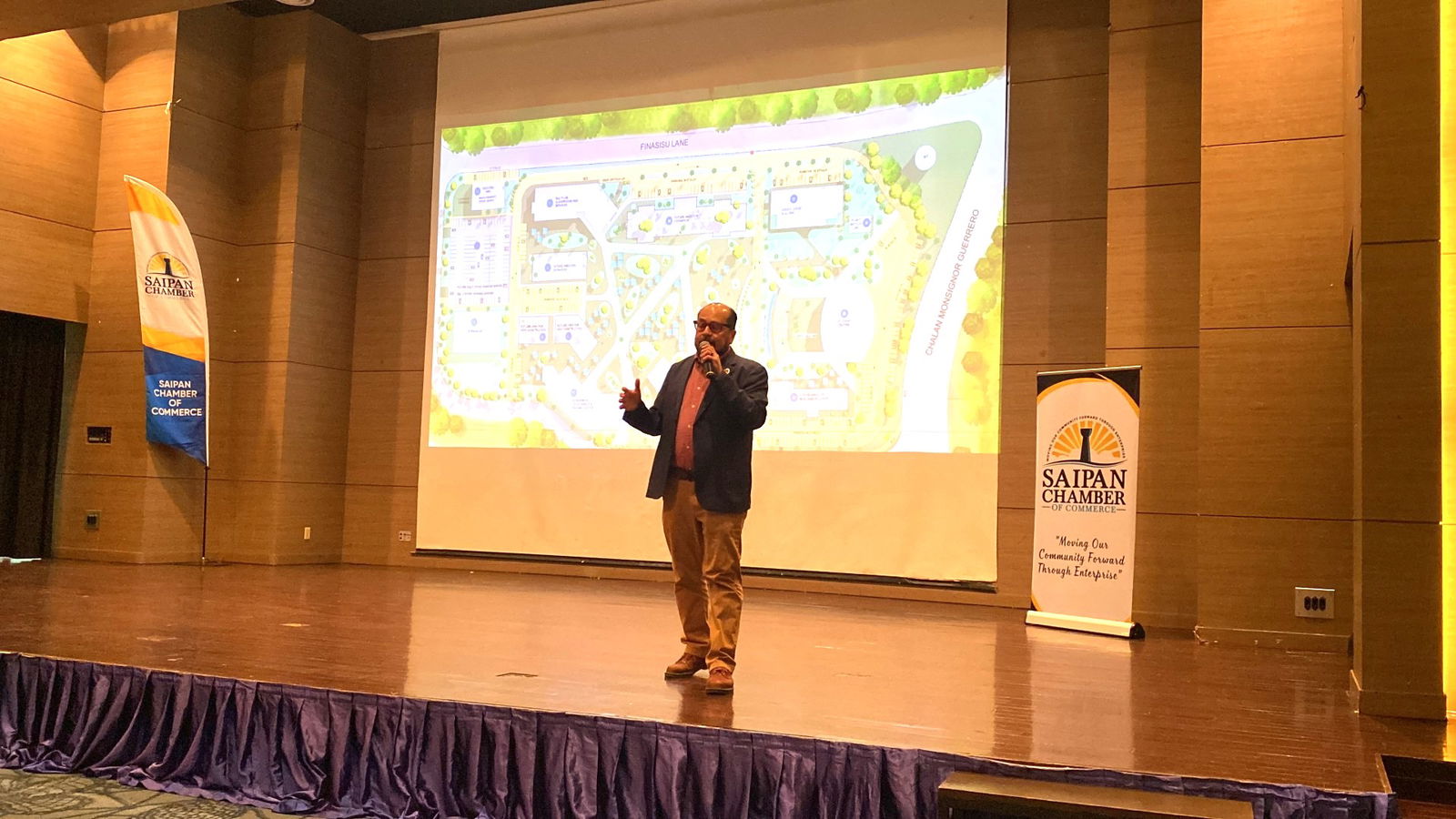 Northern Marianas College President Galvin Deleon Guerrero, EdD speaks at the July general membership meeting of the Saipan Chamber of Commerce. Behind him is a photo of the facilities master plan projects coming down the construction pipeline.